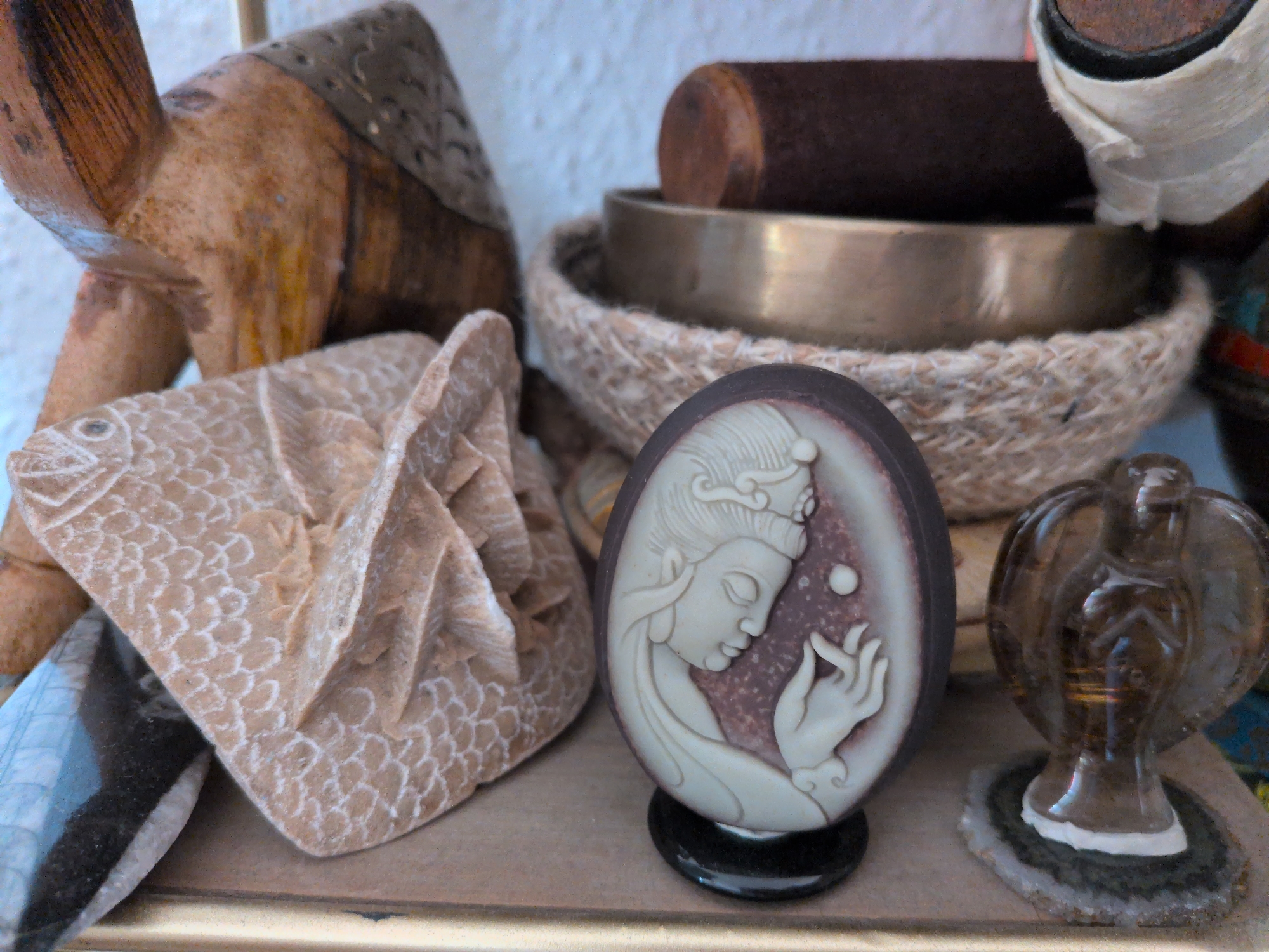 In the city of Brandenburg an der Havel, Germany, a still life arrangement of various decorative objects is displayed. The scene includes a carved wooden camel, a carved stone fish, an oval cameo with a relief of a woman's profile, a bronze singing bowl with a mallet, and a small, translucent brown glass figurine that appears to be an eagle or similar bird. The objects are arranged on a surface, likely a shelf or table, with a textured white wall in the background. The lighting suggests an indoor setting, possibly a shop or a private collection.