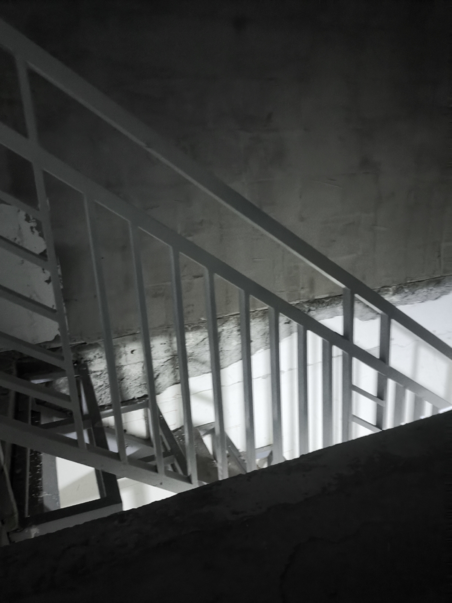 An indoor staircase made of white metal railings and concrete steps is visible. The staircase is dimly lit, with shadows cast by the railings and the structure itself. The walls are rough, unpainted concrete, suggesting an unfinished or industrial setting. The overall impression is one of a utilitarian and somewhat stark environment.