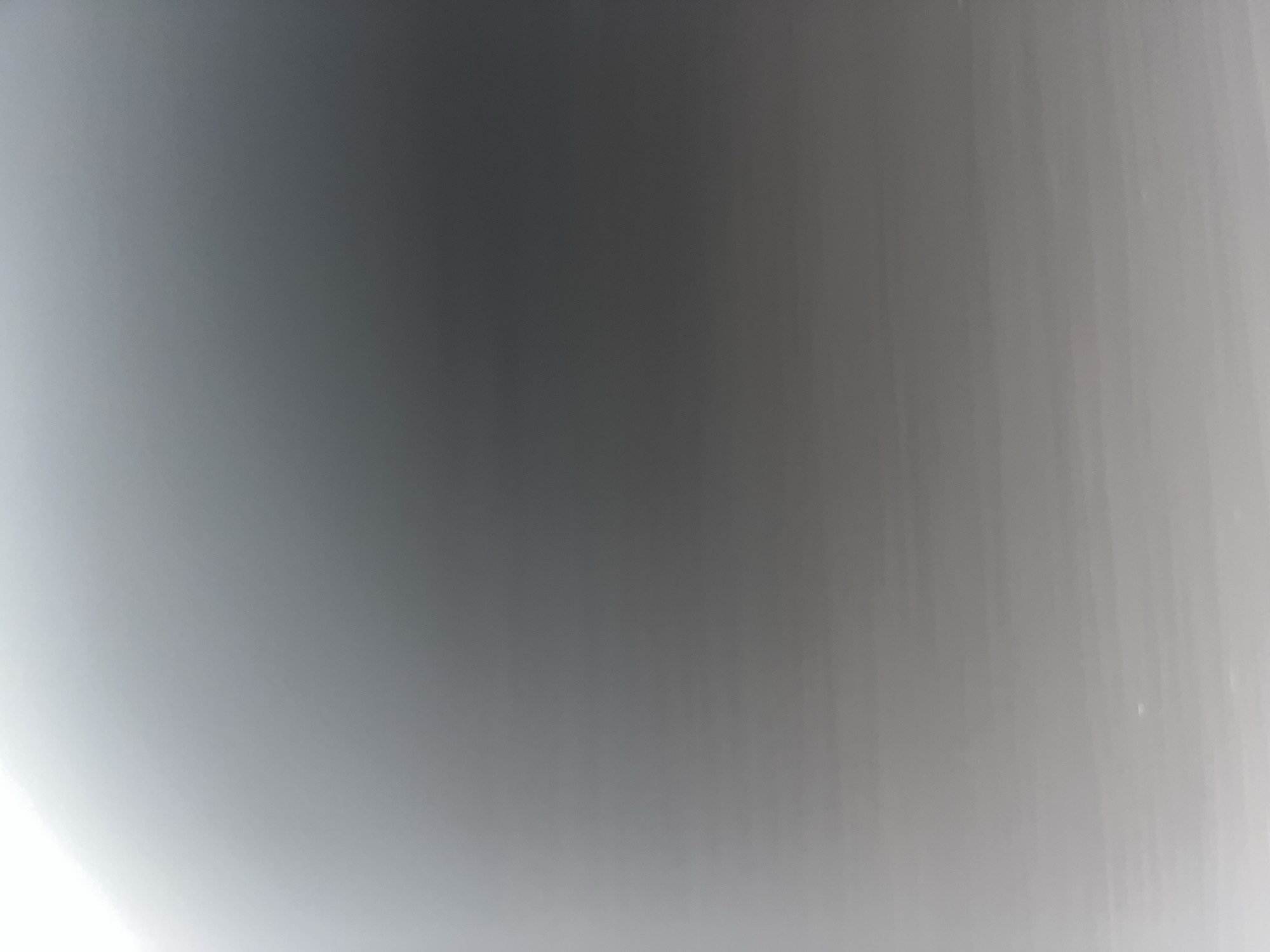 The image is a very blurry, out-of-focus shot of a light gray surface that appears to be a ceiling or a wall. There are subtle horizontal lines or streaks visible in the lower portion of the image, suggesting a textured surface or possibly a light reflection. The lighting is diffuse, with a brighter area in the upper left corner that gradually darkens towards the bottom right. There are no discernible objects, people, or actions depicted in the image, and therefore no context about the location can be provided.