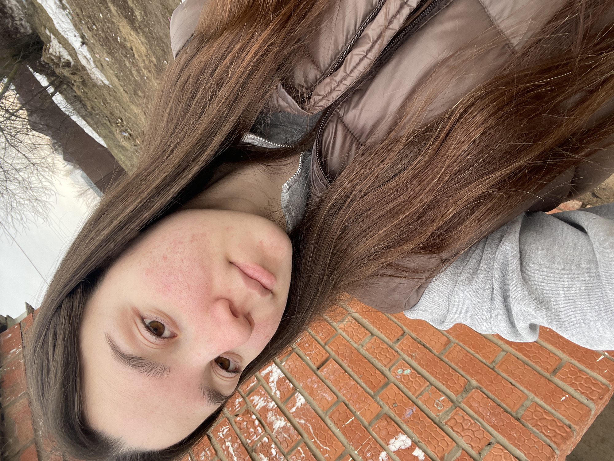 A young woman with fair skin, brown eyes, and long brown hair is centered in the image, angled towards the camera. She is wearing a brown puffer vest over a gray sweatshirt. The background shows a brick surface with patches of white, possibly snow, and a blurred outdoor scene with bare trees. The photo appears to be taken in Ольховка, Russia, given the instructions.