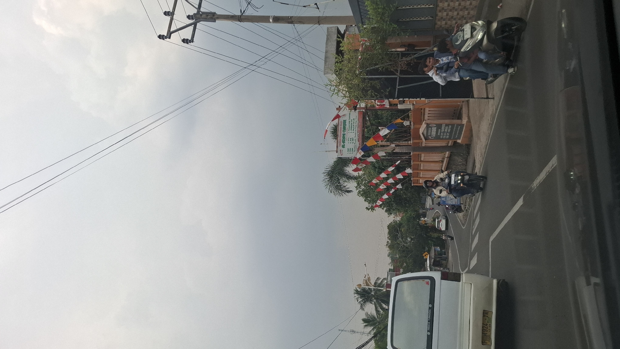 The image depicts a street scene in Sukasari, Indonesia, under an overcast sky. The foreground shows the rear of a white van with a license plate "F 1924 YR 12-26". To the right, a paved road curves, with a pedestrian crossing marked by white lines. A man wearing a light-colored jacket and sunglasses rides a dark-colored motorcycle in the middle lane, facing away from the camera. Further down the road, three young men are riding on another dark-colored motorcycle, with the driver in front wearing a light blue jacket.
On the right side of the road, there's a building with a sign that appears to be an educational institution, possibly a school, as indicated by the text "AL-AKSAR CENDIKIA" and a list of educational levels such as "1. SMP", "2. SMA", "3. SMK". This building is adorned with red and white Indonesian flags, also known as "pennants" or "banderoles", which are common during national celebrations. Trees, including palm trees, are visible behind the building. Electrical poles and wires are present in the background, typical of urban settings. The overall atmosphere suggests a day with limited sunlight.