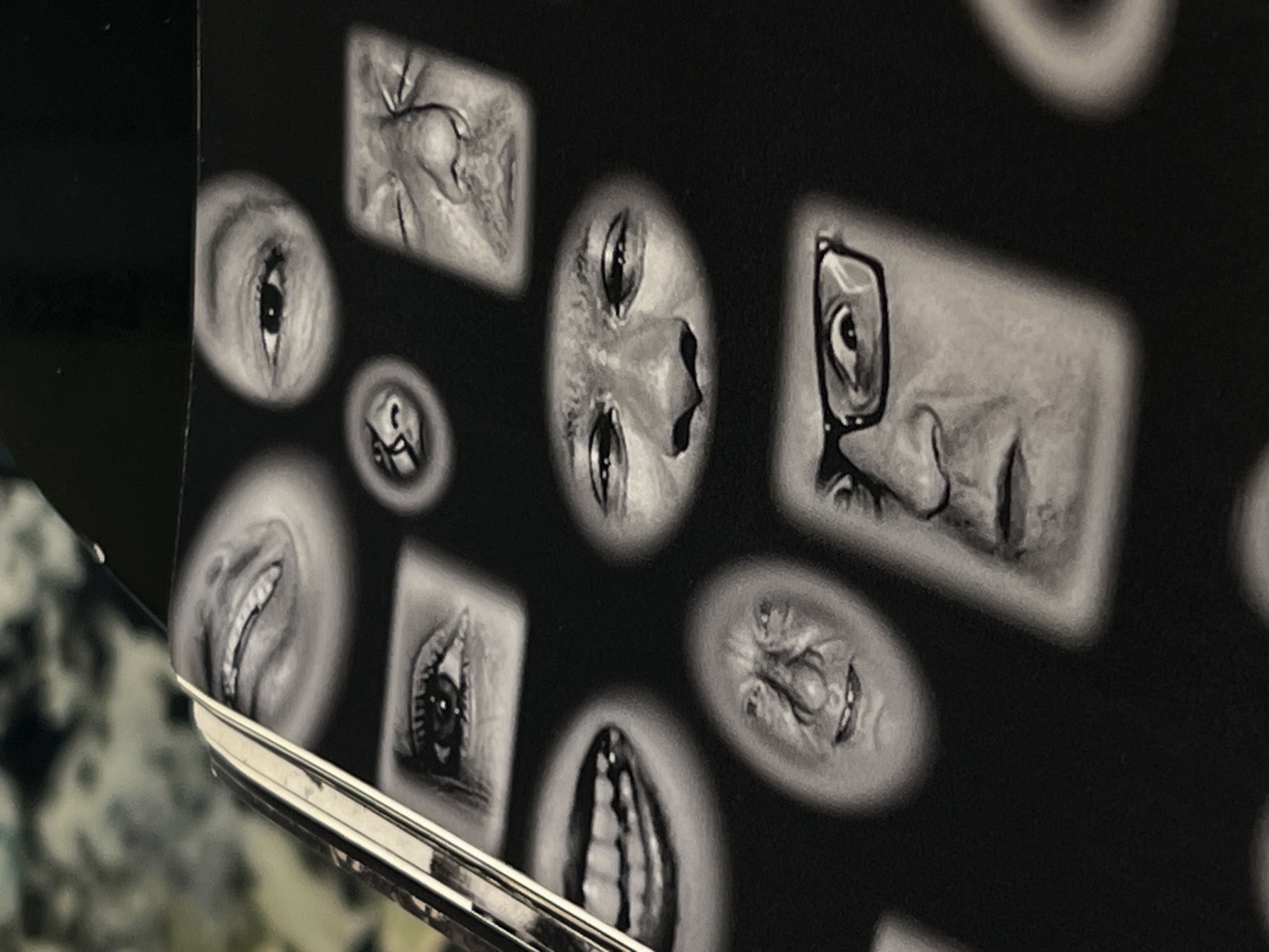 The image is a close-up of a black and white surface adorned with surreal, fragmented drawings of facial features, including eyes, noses, and mouths. One drawing depicts a person wearing glasses. The metallic edge of an object, possibly a book or device, is visible at the bottom left corner of the frame. The background is a blurry, out-of-focus area with a hint of a patterned texture. There is no indication in the photo suggesting this image was taken in Tel-Aviv, Israel.