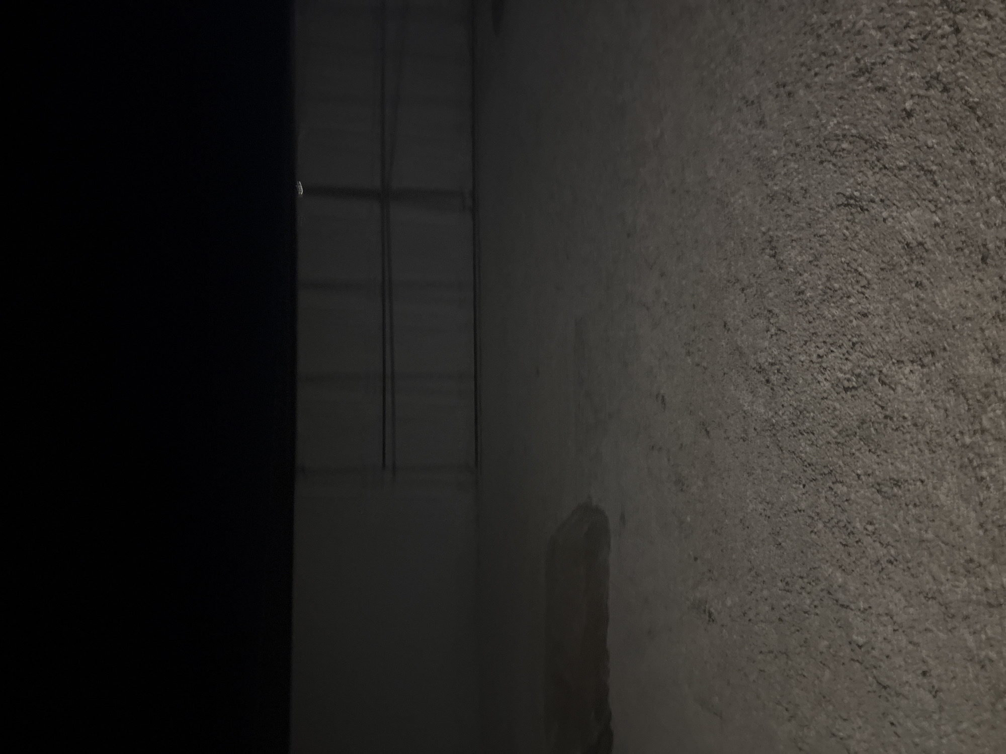 The image depicts a dimly lit, outdoor scene. In the foreground, the ground appears to be rough asphalt or gravel. A dark, oblong object, possibly a large piece of cloth or debris, lies on the ground to the left. In the midground, there is a large, light-colored wall or structure, with horizontal lines suggesting panels or doors. The upper portion of the image is entirely black, indicating darkness or a very dark sky. The overall impression is of an industrial or utilitarian area at night, with no clear indication of its specific location within Burgas, Bulgaria, beyond it being an outdoor, possibly abandoned or service area. There are no people or animals visible in the frame.