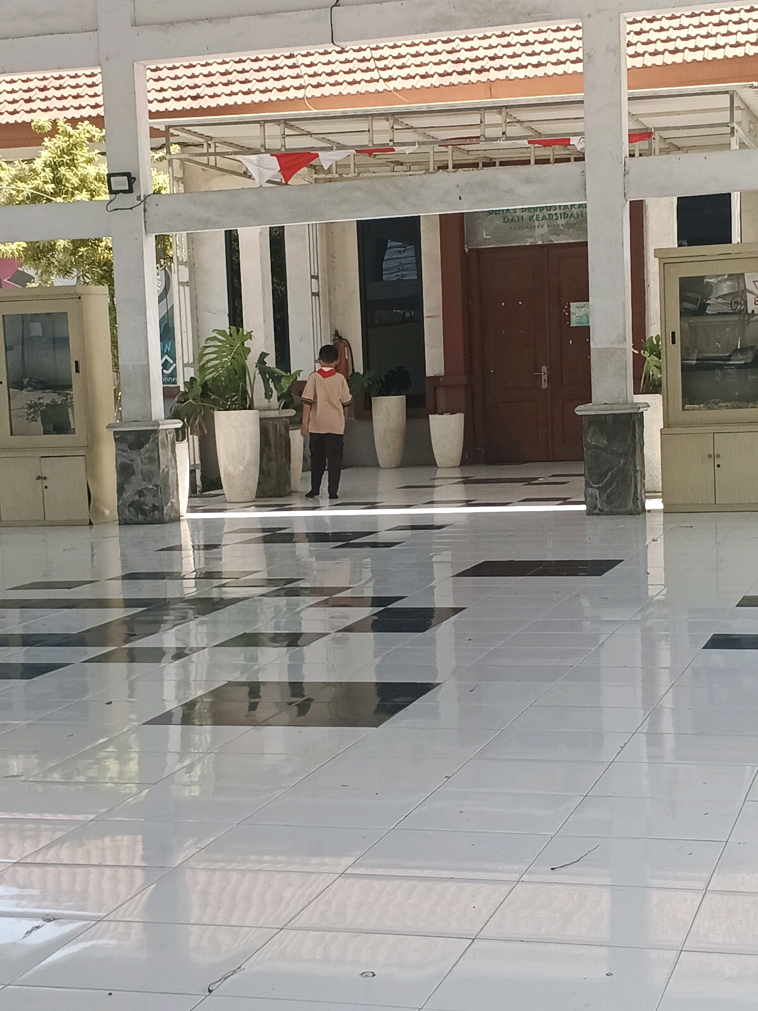A young boy wearing a light brown shirt and dark pants with a red collar stands in the middle of a tiled floor, facing away from the camera. He is in front of a building with large brown double doors and a sign above them. The building appears to be a public institution, possibly a library or government office, as indicated by the sign which reads "DINAS PERPUSTAKAAN DAN KEARSIPAN KABUPATEN SIDAN". The floor is tiled in a black and white checkered pattern, which is highly reflective, creating mirror-like images of the surroundings. The area is an open walkway or veranda, with pillars and a roof overhead. Large potted plants are placed around the entrance. In the background, the roof of the building has red tiles, and flags, possibly red and white, are strung across the upper structure. The scene appears to be in Krian, Indonesia, based on the sign.