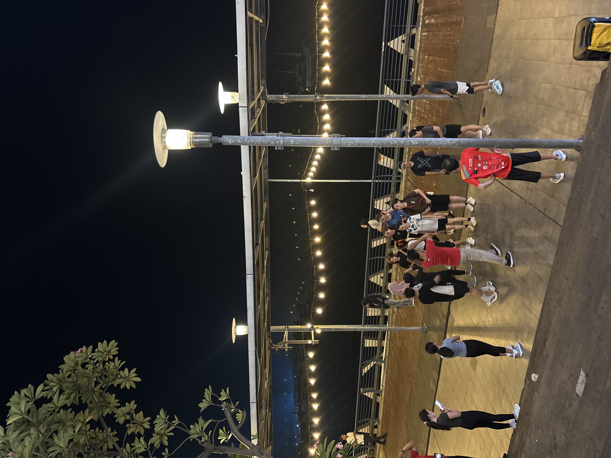 A nighttime exterior shot captures a group of approximately 15 to 20 individuals gathered on a paved walkway beside a body of water. The walkway is illuminated by overhead lights and a string of decorative bulbs stretched across the scene. A large tree with broad leaves is visible on the left side of the frame, with a few pink flowers. In the background, lights from buildings and several boats are visible on the water. The people are dressed casually in athletic wear, and some appear to be engaged in activities or conversations. The overall atmosphere suggests an outdoor public space, possibly a waterfront promenade or park in North Jakarta, Indonesia.