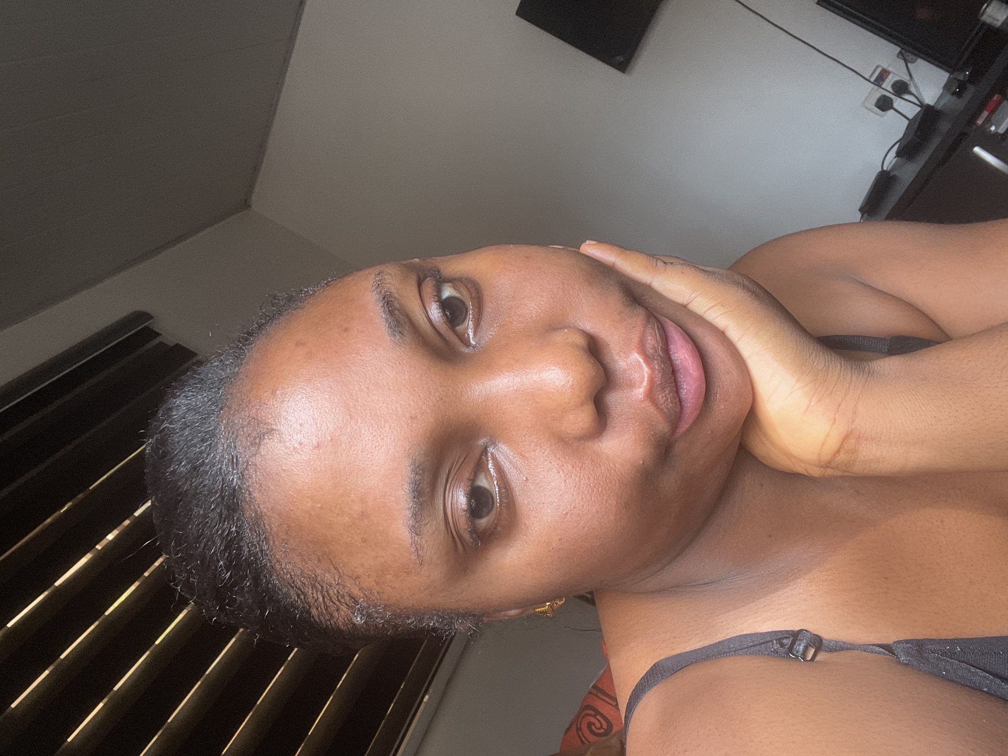 A close-up selfie of a Black woman with short, dark hair, lying down, with her head tilted to the left. Her left hand is touching the side of her face. She is wearing a black tank top. The background shows a bedroom with a window with blinds and a wall with a television and cords. The location of the photograph is potentially in Abuja, Nigeria.