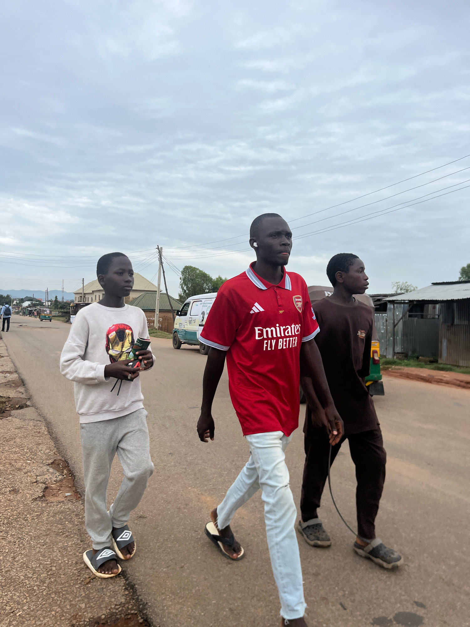 Three young men are walking on a paved road in Barakin Akawo, Nigeria. The man in the center is wearing a red Arsenal soccer jersey with "Emirates Fly Better" printed on it, white jeans, and dark slide sandals. He has white earbuds in his ears. The man to his left is wearing a light-colored crewneck sweatshirt with a graphic design of a helmet, gray sweatpants, and black and white slides. He is holding what appears to be a packaged item. The man to the right is wearing a dark brown t-shirt and dark pants, with dark clogs on his feet. He is holding a dark cord that trails behind him. The road is lined with sparse vegetation and buildings, including corrugated metal structures and a white van. The sky is overcast.