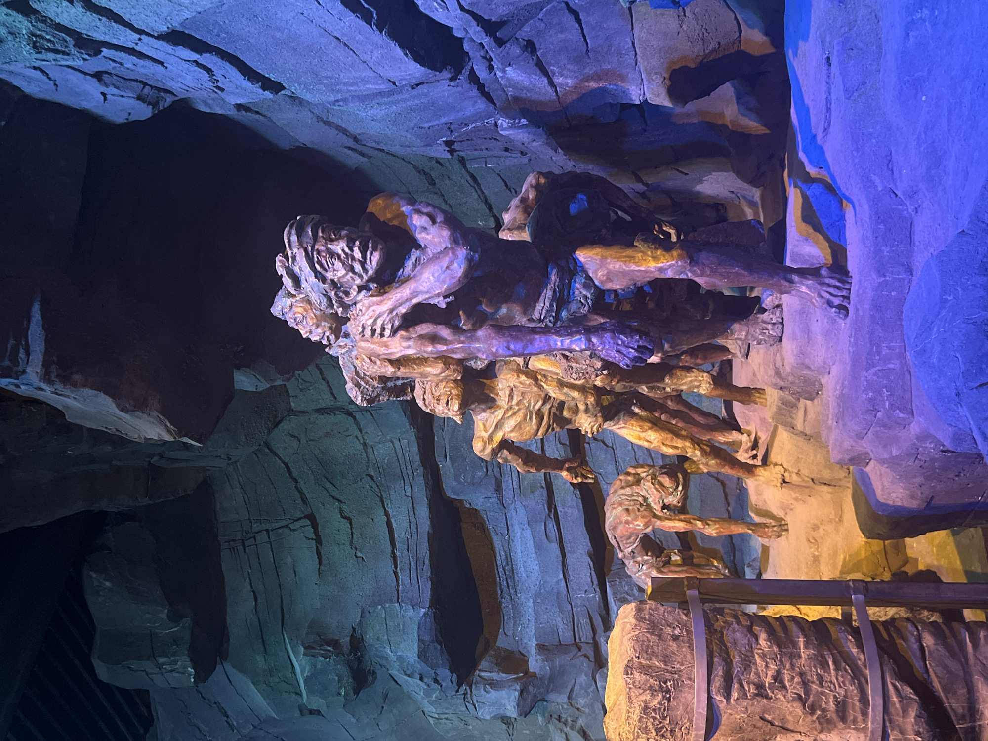 The image depicts a dramatic scene within a cavernous, rocky environment, likely a theatrical set or an attraction. At the center, a group of figures, appearing to be statues or mannequins, are posed in strenuous activity. The primary figure, a muscular male, carries a heavy object or another person on his shoulder, his face contorted in exertion. Behind him, several other figures are similarly depicted in poses of struggle and labor, their bodies strained and their expressions conveying effort. The figures are rendered in a metallic or bronze-like material, with areas highlighted in gold. The environment is characterized by rough, textured rock formations that surround the figures. The lighting is dramatic, with strong blue and purple hues illuminating the scene from the right, casting deep shadows, while a warmer, orange light bathes the left side of the foreground. The background features dark, cave-like openings and the ceiling of the structure appears to be made of dark beams or panels. The image is a close-up shot, focusing on the sculptures and their immediate surroundings, and does not provide any explicit information about a specific location in Yuzhong District, China, or any other identifiable geographic context beyond the sculpted scene itself.