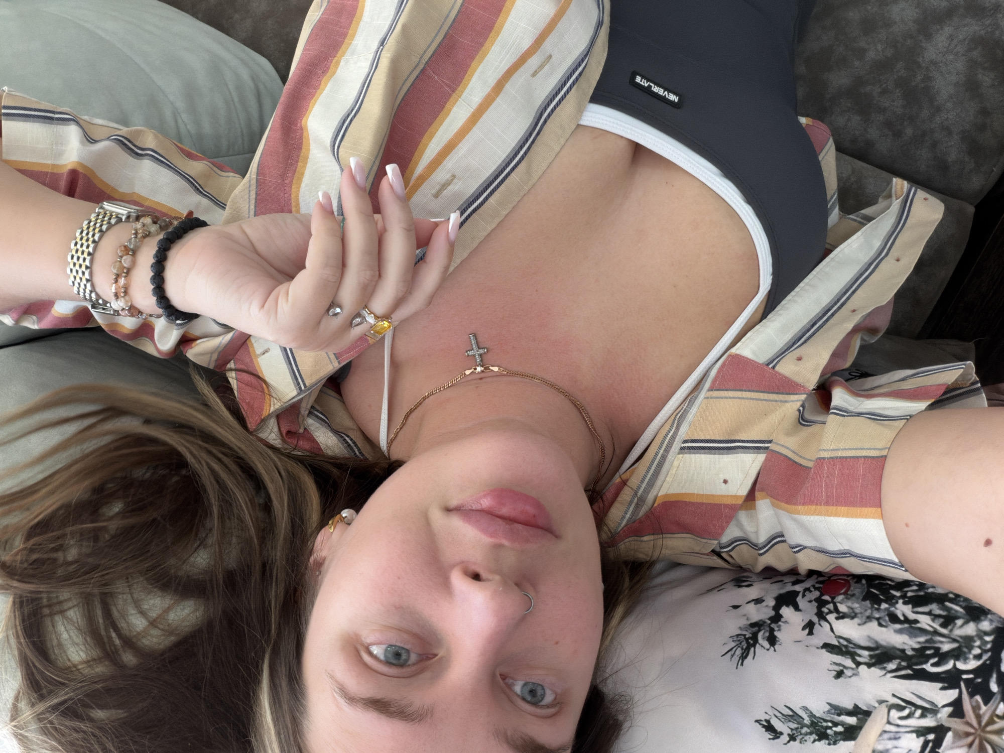 The image presents an overhead, close-up view of a person, likely a woman, lying down indoors, taking a selfie. Her face, positioned towards the bottom of the frame, features blue eyes, light brown hair, and a silver septum piercing. Her lips are slightly parted, and her expression is neutral as she gazes directly at the camera.
She is dressed in a loose, open button-up shirt characterized by multi-colored stripes in hues of beige, red, orange, and blue. Beneath this, a dark grey sports bra or crop top with white trim is visible, prominently displaying the white text "NEVERLATE" across the chest.
The person wears several pieces of jewelry. Her left wrist is adorned with a silver-toned watch and two beaded bracelets, one light-colored and one dark. Multiple rings, including one with a distinct yellow stone, are worn on her fingers, which have French-manicured nails. Around her neck, two necklaces are layered: one is a delicate gold chain with a small, sparkly cross pendant, and the other has a small, heart-shaped clasp at the back. A gold-colored earring with a white pearl is visible in her right ear.
She appears to be reclining on soft furnishings, possibly a couch or bed. Her hair is spread out on a plain light green cushion. Part of another pillow, featuring a white, green, and black leafy pattern, is visible towards the right. The setting suggests a comfortable, domestic indoor environment. No specific time of day or external location cues are discernible.