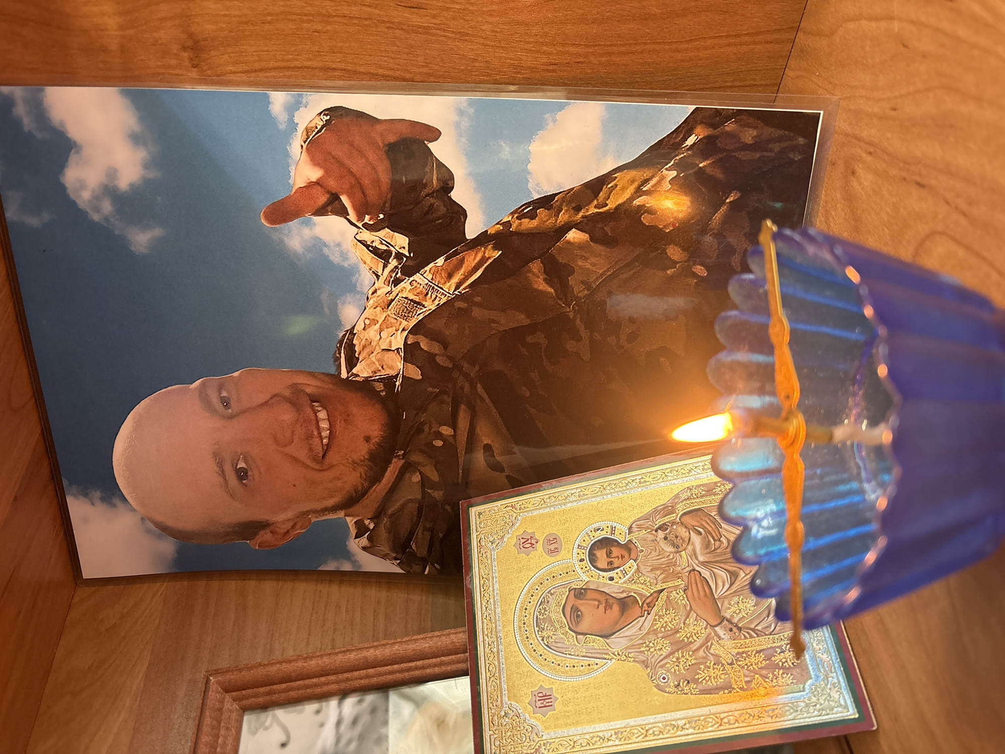 A photograph shows a collection of items placed on a wooden surface, possibly a shelf or cabinet. A photograph of a man in military camouflage fatigues is displayed prominently. The man is smiling and pointing, with a background of sky and clouds. To the right of the man's picture is an Orthodox icon of the Virgin Mary holding the child Jesus. A blue glass candle holder with a lit candle stands in front of the icon. The surface and background are made of a light-colored wood. There is no indication in the image regarding the location in the city of Perm, Russia.