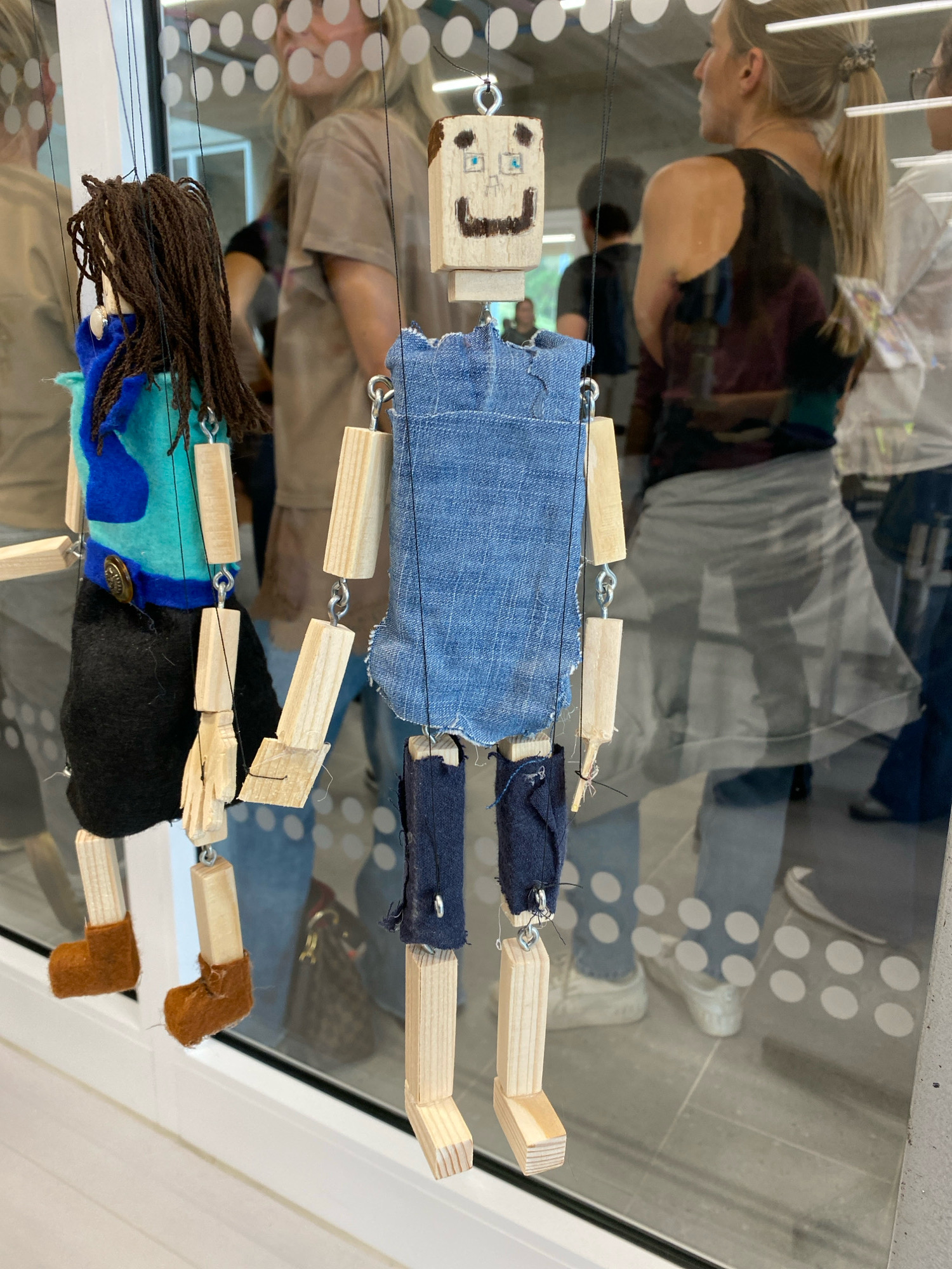 The image shows two handmade wooden marionette puppets hanging in front of a large window. The puppet on the left has brown braided hair, a blue and green top, a black skirt, and brown felt boots. The puppet on the right has a wooden head with a drawn-on face, a blue denim tunic, and dark blue shorts. Both puppets are constructed from wooden blocks connected by metal chains. In the background, through the window, blurred reflections of people can be seen, along with the interior of what appears to be a public building or exhibition space. The floor inside has a pattern of white circles. The window is framed by a white sill and vertical metal supports. There is no visible indication of the location being in Horhausen (Westerwald), Germany.
