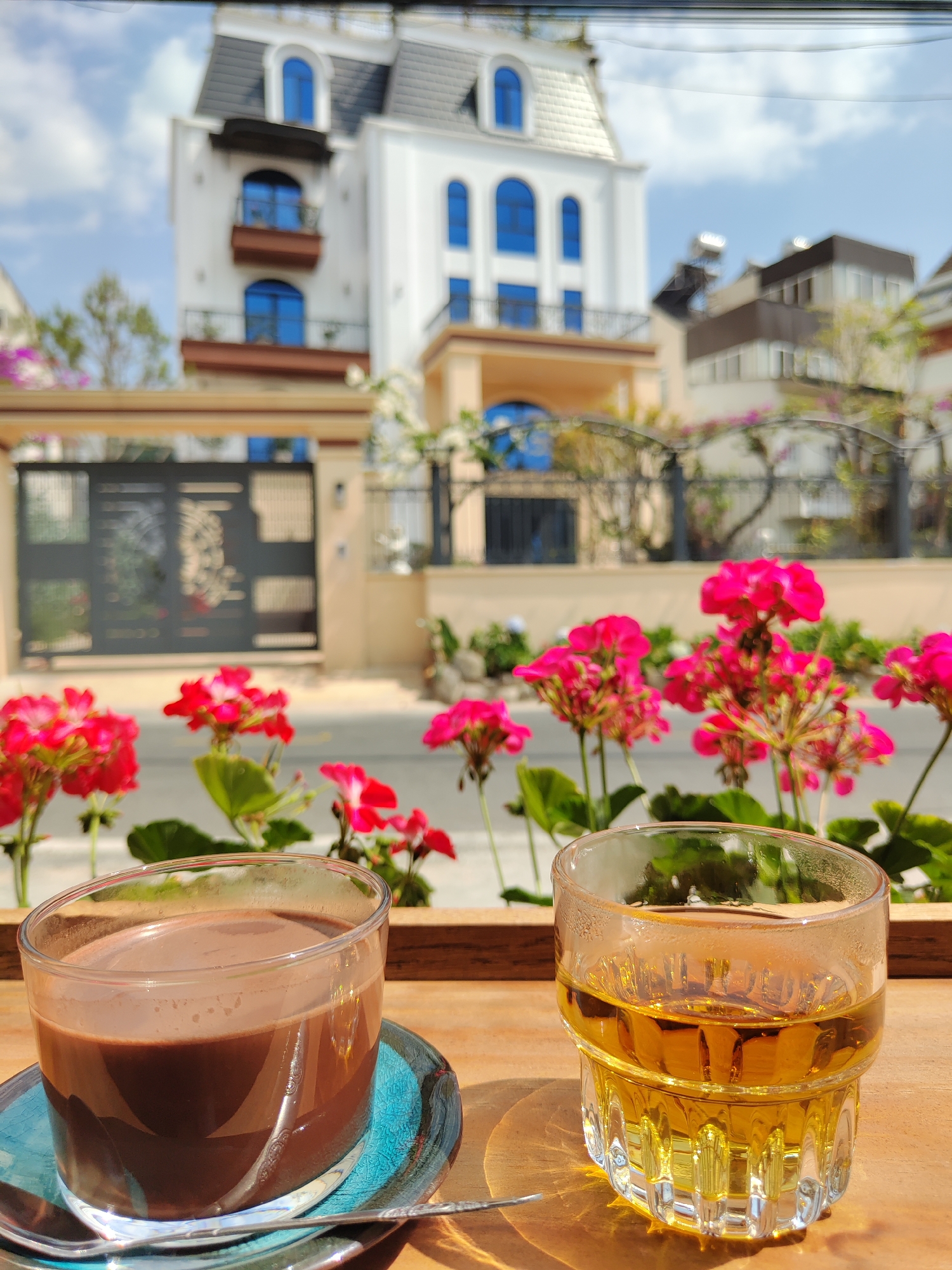 The photograph captures two beverages, one chocolate and one a clear golden liquid (tea), placed on a wooden surface. Pink geraniums are visible in the midground. In the blurred background, a white building with distinctive blue-framed arched windows stands prominently, along with a gate and other buildings. The location is Lâm Viên - Đà Lạt, Vietnam.
