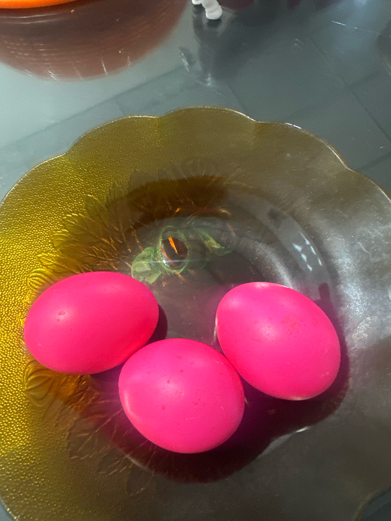 Three brightly colored pink eggs are placed in a textured, iridescent brown glass bowl. The bowl has a scalloped edge and a raised pattern on its surface. The eggs are oval and appear to be hard-boiled, with smooth shells. The bowl sits on a reflective, dark surface, possibly a table. In the background, out of focus, are blurry objects that suggest an indoor setting, potentially a kitchen or dining area. There is no indication of the specific location within the City of Binjai, Indonesia, as no discernible landmarks or identifying features are present.