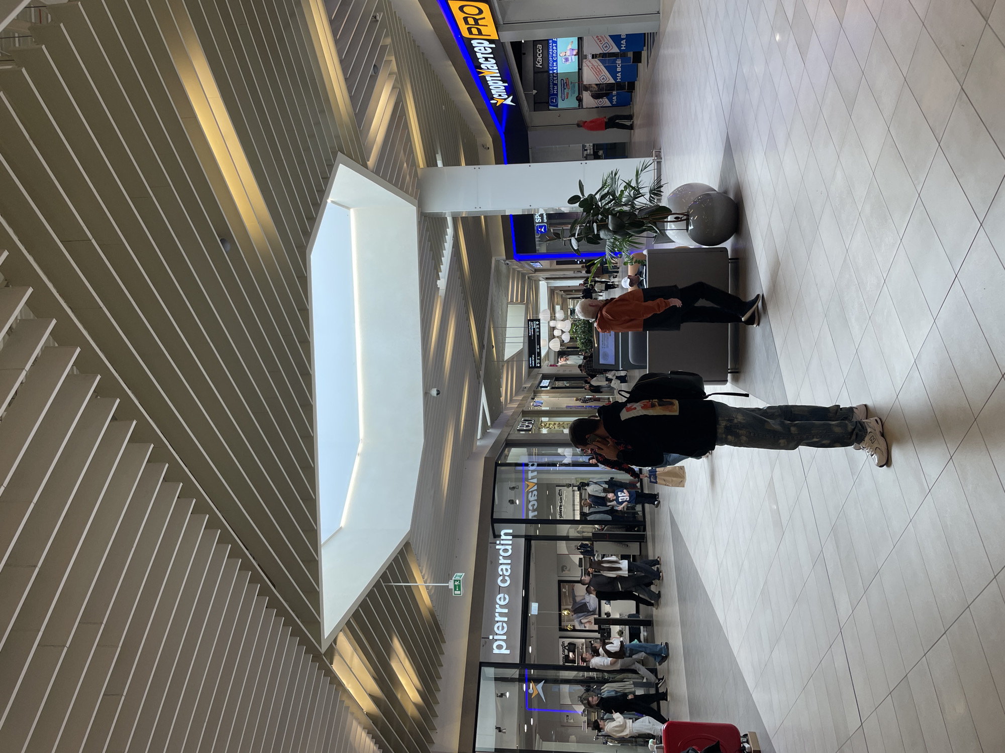 The image shows an interior view of a shopping mall, likely in Kazan, Russia, due to the Cyrillic text. The mall has a high, architecturally interesting ceiling with a large rectangular skylight. Below the skylight, the ceiling consists of closely spaced, parallel beams with integrated lighting. The floor is tiled in a light color.
Along the right side of the image, there are seating areas with gray rounded benches and potted plants. A man with a backpack is standing in the aisle, looking at a store. In the background, another person is sitting on one of the benches.
On the left side of the image, there is a Pierre Cardin store. Shoppers and shop assistants are visible through the store's glass facade. Further down the corridor, a "Sportmaster PRO" store is visible with its sign in both Cyrillic and English.
The scene suggests a modern shopping environment with a focus on design and natural light. The presence of people implies active commerce and customer activity. A red luggage is at the bottom of the frame.
