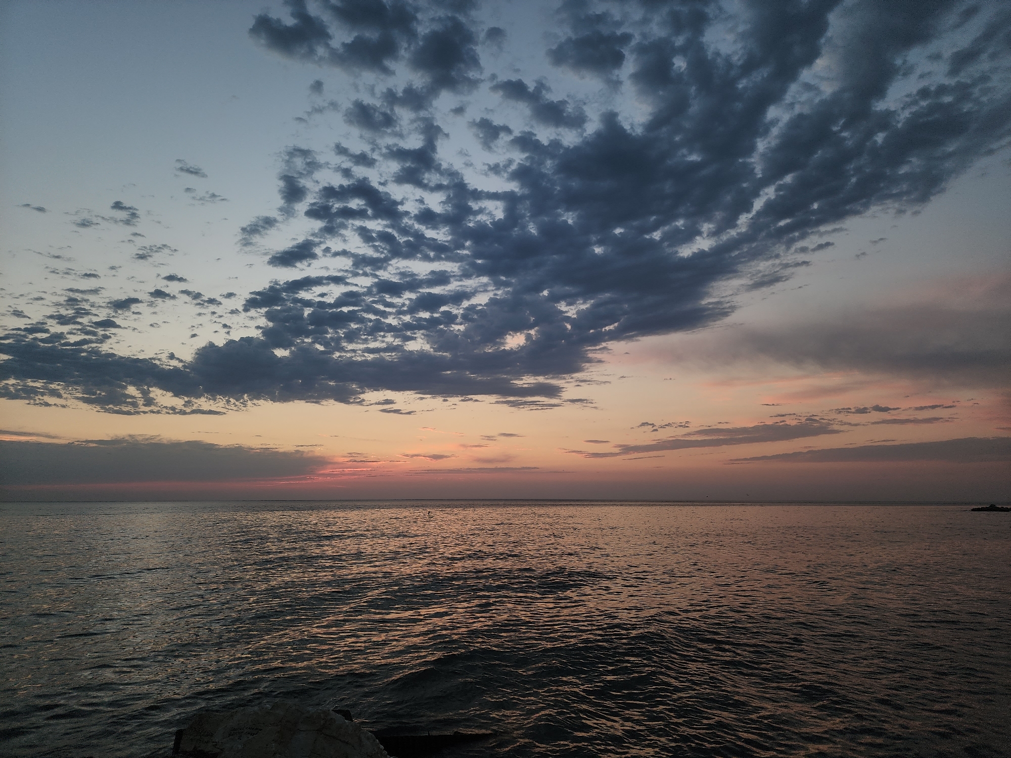 The image depicts a serene seascape at dusk, likely near Makhachkala, Russia. The sky is a gradient of soft blues and grays, transitioning to warm hues of pink and orange near the horizon, indicating the setting sun. Wispy clouds are scattered across the upper portion of the sky, with darker, more prominent clouds dominating the upper left. The sea is calm, with gentle ripples reflecting the muted colors of the sky. A rocky formation is visible in the lower left foreground, partially submerged. The horizon line is clear, separating the sea from the sky. In the far distance, on the right side of the horizon, a faint dark shape, possibly a buoy or a distant structure, is discernible. The overall atmosphere is tranquil and contemplative.