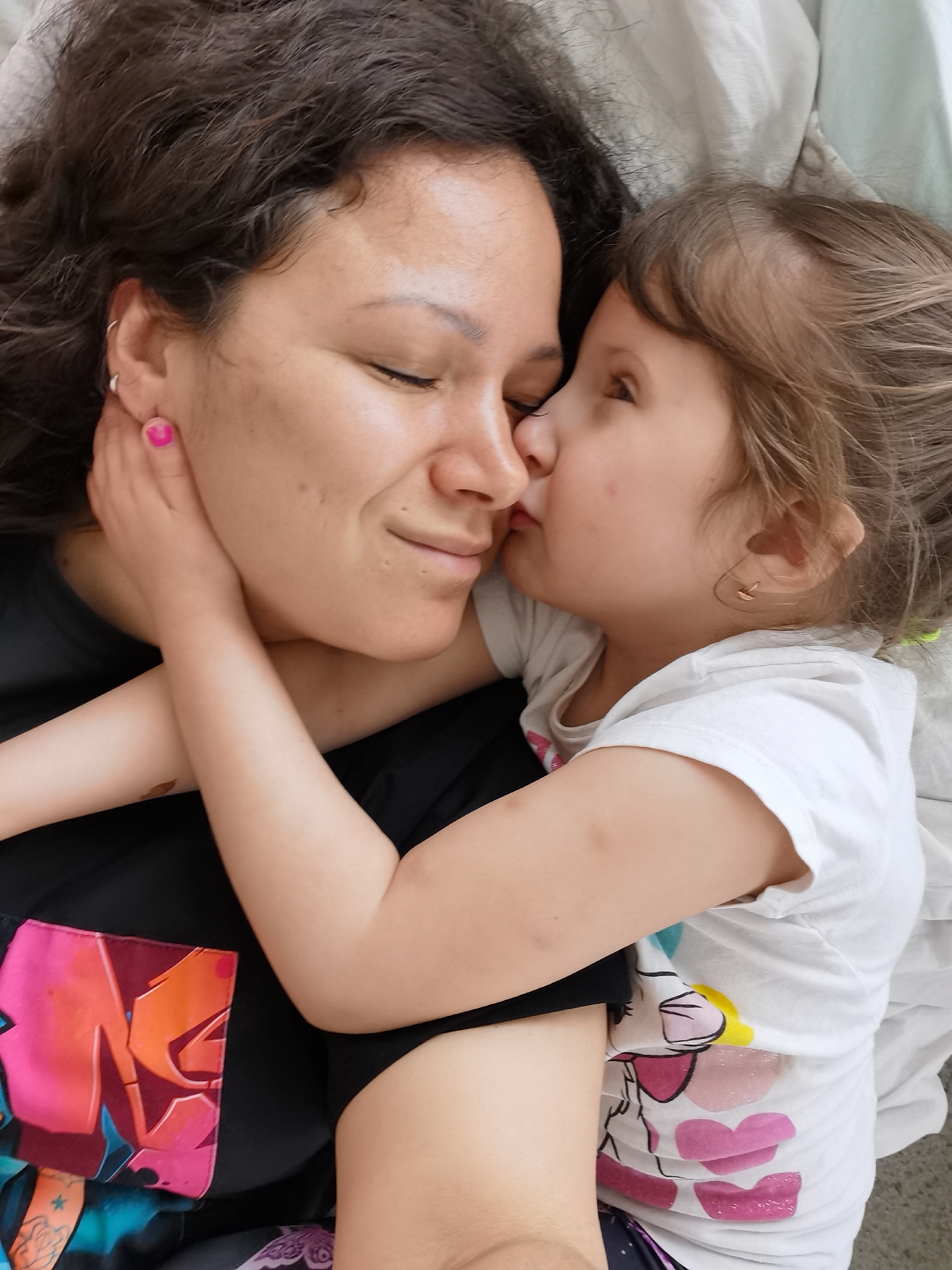 A woman and a young child are lying down, likely on a bed, indoors. The woman has dark, curly hair and is wearing a black t-shirt. The child, with lighter brown hair, is nestled close, with an arm around the woman's neck and is kissing her face. The child is wearing a white t-shirt. The location in the city of Perm, Russia cannot be determined as it is not visible in the image.