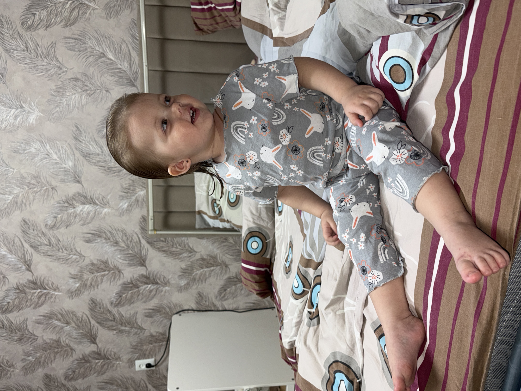 A young child lies on a bed with patterned bedding in a room with feather-patterned wallpaper. The child is wearing grey pajamas with a bunny and floral pattern. A mirror and a white object, possibly a table, are visible in the background. The location is Reshetnikovo, Russia.