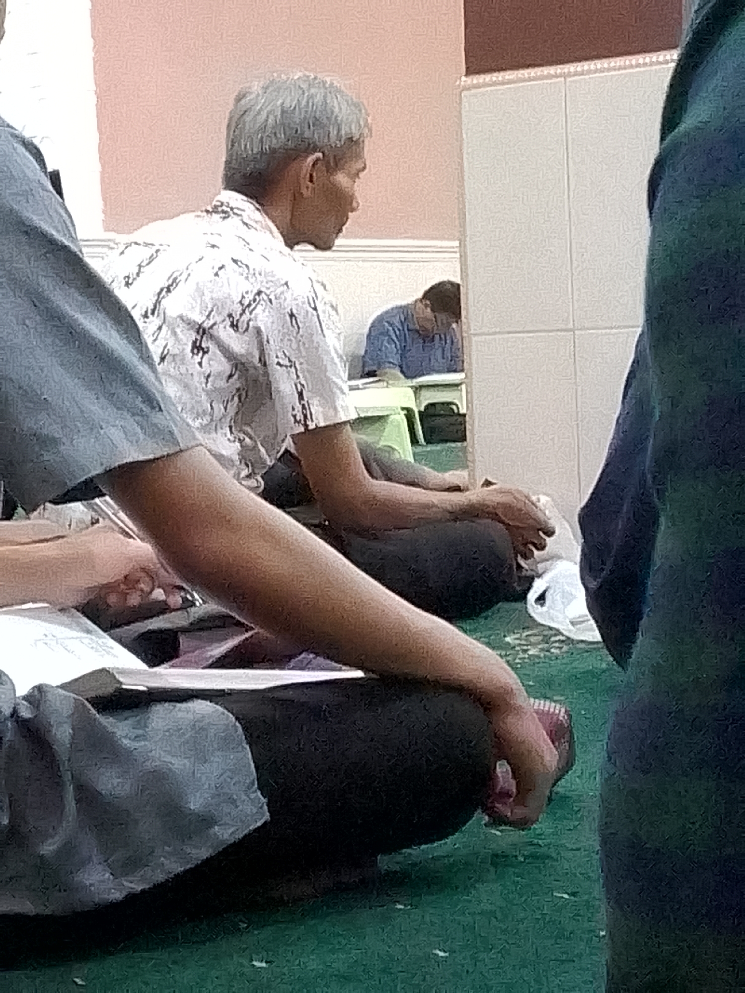 The image shows an indoor scene, likely a mosque or prayer hall, with several people seated on a green carpeted floor. In the foreground, a person in a blue shirt is partially visible on the left, with their arm resting on an open book. To their right, an older man with gray hair wears a white shirt with a black pattern and is also seated on the floor, his hands resting on his lap. Behind him, another individual in a blue shirt is seated and appears to be looking down. The background consists of a wall with tiled sections. The environment suggests a place of worship or study, potentially in the city of Kertajaya, Indonesia, given the context provided.