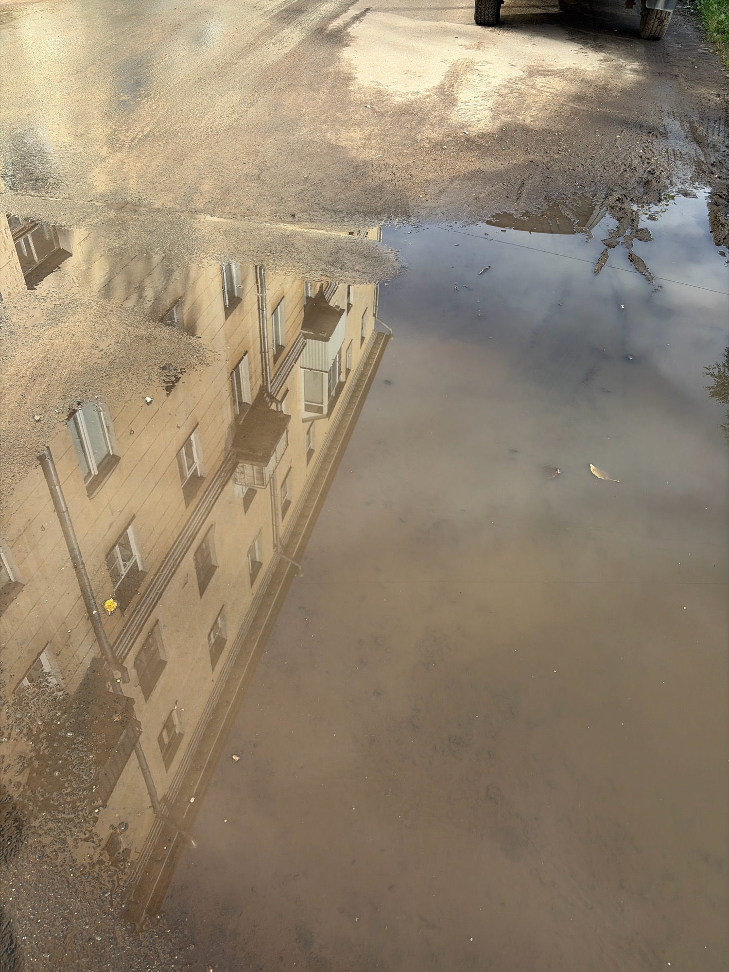 The image shows a muddy puddle reflecting a multi-story building with light-colored walls and numerous windows. The building appears to be residential. The reflection is distorted by the water's surface and the muddy content of the puddle. The ground around the puddle is a mix of asphalt, dirt, and gravel. A portion of a vehicle with tires is visible in the upper right corner of the image, suggesting it is parked or driving on the road. The overall scene depicts an urban environment, likely in a city like Arkhangelsk, Russia, after rain or during a period of wet weather.
