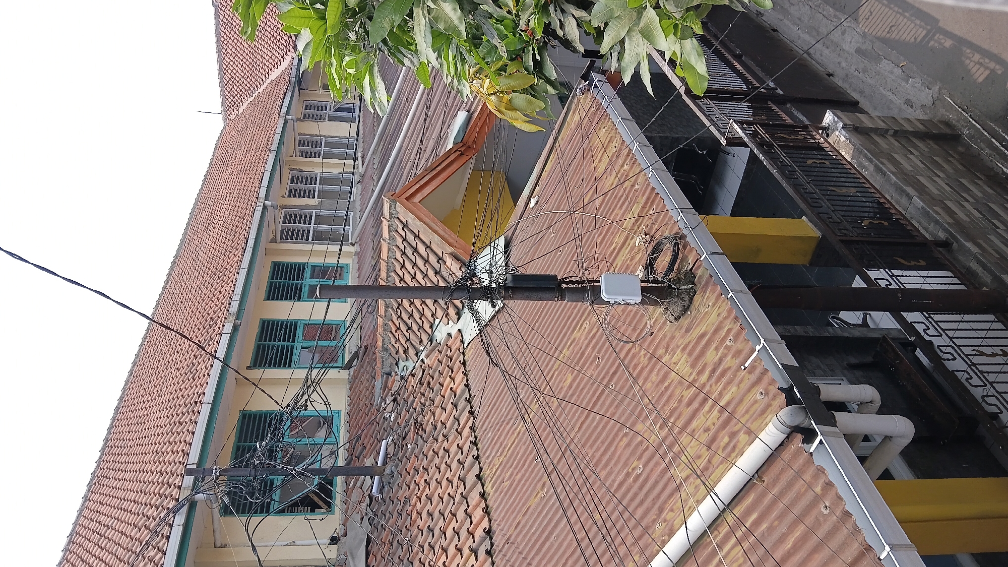 The image depicts a cluster of residential buildings with red tile roofs and yellow walls, characterized by an abundance of overhead electrical and telecommunications wires. A prominent utility pole, cluttered with wires and several electronic boxes, stands in the foreground, rising above the rooftops. The architecture suggests a densely populated urban environment. The presence of multiple windows with teal-colored frames on the upper building indicates living spaces. The lower section of the image shows a fence and the exterior of a building with a balcony. A large green tree is visible on the right side, partially obscuring the view. The scene appears to be an outdoor view from an elevated position, possibly a higher floor or adjacent building. The setting is consistent with a residential area in a city like Bandung, Indonesia, where such dense wiring is common.