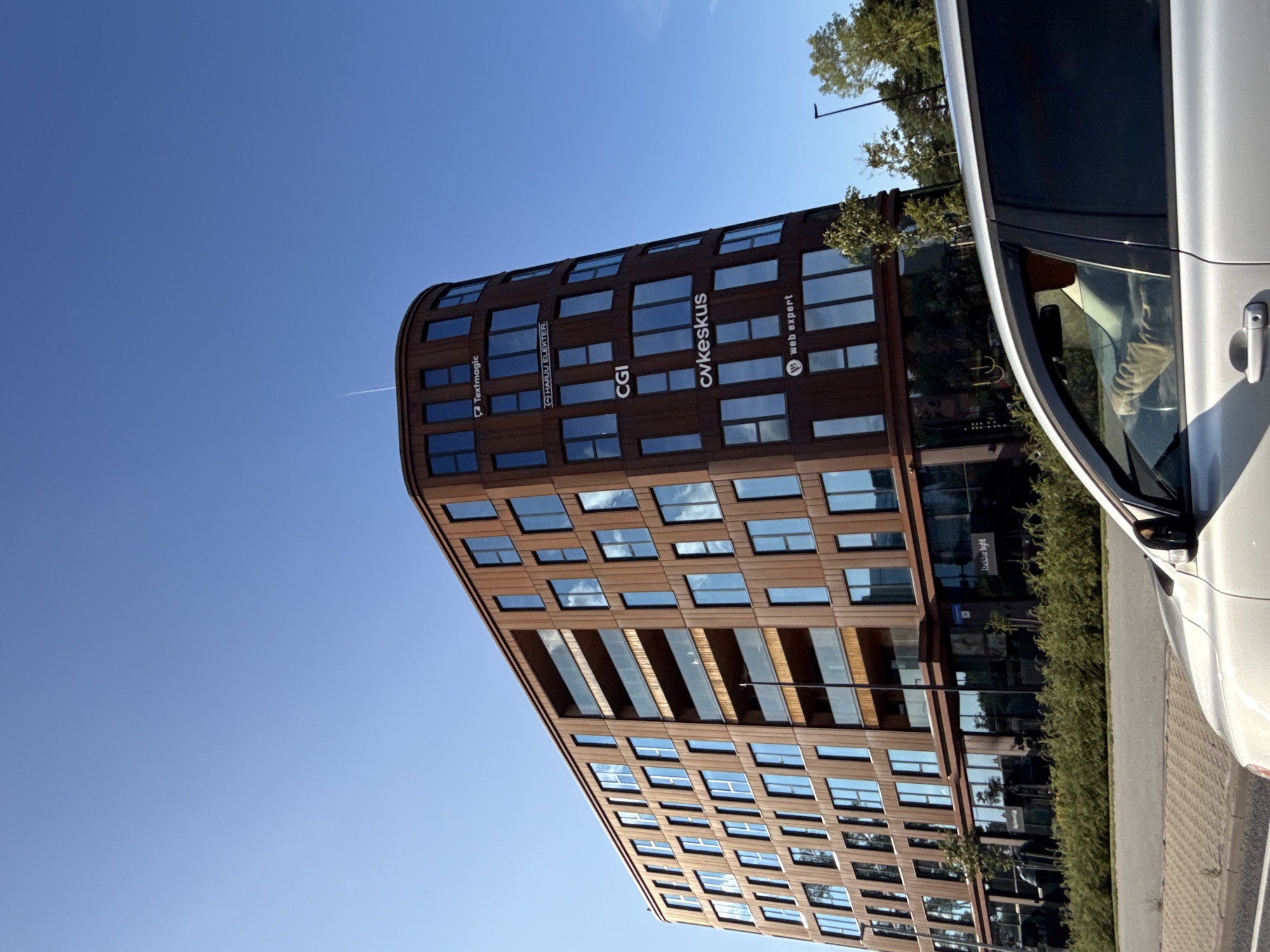A modern, curved building with a brown, metallic facade and large windows dominates the frame under a clear blue sky. Several company logos are visible on the building's exterior, including "Textmagic," "HARJU ELEKTER," "CGI," "cvkeskus," and "web expert." The building appears to be located in the city of Tallinn, Estonia. In the foreground, the top of a silver car is visible, with a person's hands on the steering wheel. The car is positioned on a road in front of the building. Green bushes and trees are visible around the base of the building and along the roadside. The scene suggests an urban setting on a sunny day.