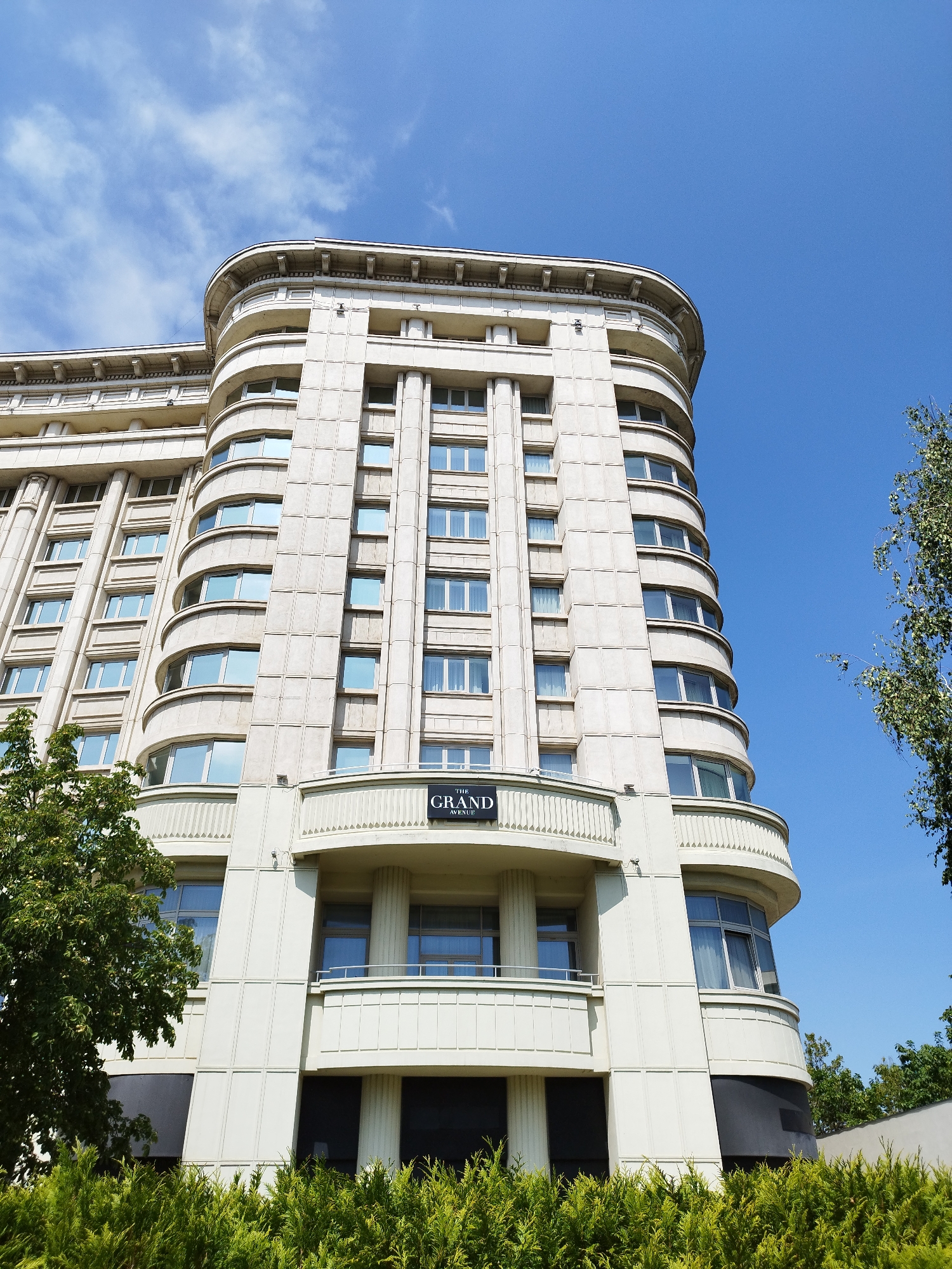 The image shows a low-angle exterior view of The Grand Hotel, located on Calea Victoriei in Bucharest, Romania. The hotel is a tall, multi-story building with a cream-colored facade and distinctive curved architecture. Numerous windows are visible, many with curtains drawn. The building features decorative architectural elements, including columns and recessed balconies. The top of the building has a rounded corner. A sign with "THE GRAND AVENUE" is visible on the building's facade. Lush green trees and shrubs are in the foreground and on the right side of the image. The sky is clear and blue with some wispy white clouds. The photograph is taken on a sunny day, with the sun casting shadows on the building.