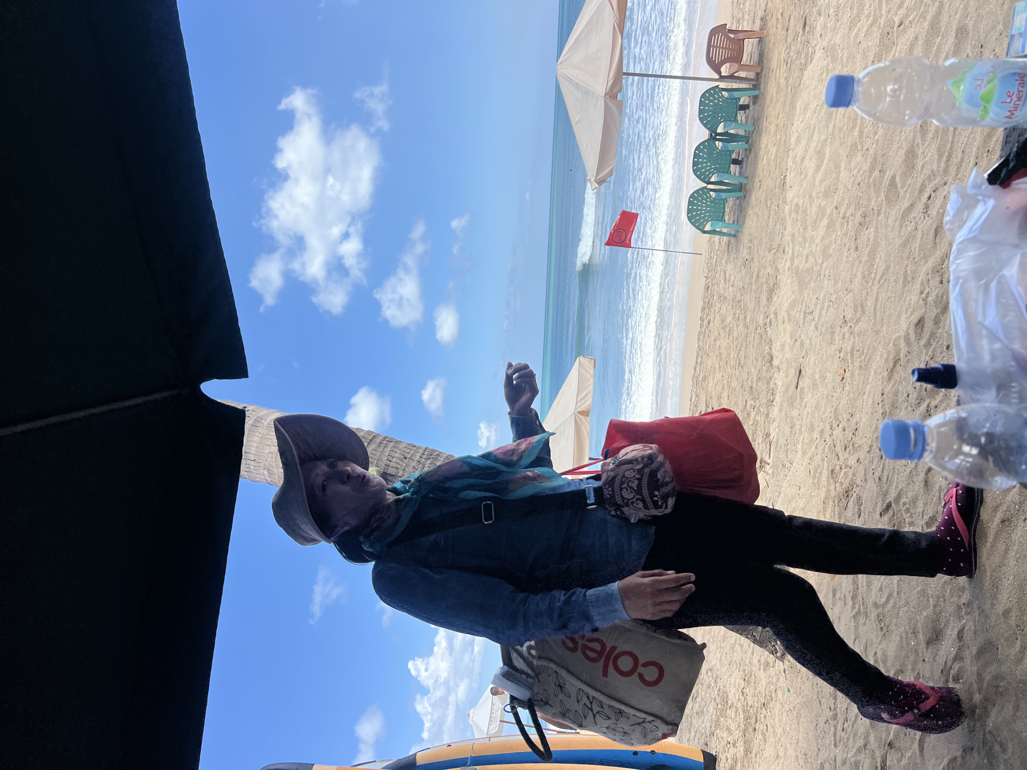 A person stands on a sandy beach in Kuta, Indonesia, under a dark awning. The person is wearing a wide-brimmed hat, a denim-like shirt, dark pants, and patterned flip-flops. They are carrying two bags: a large tote bag with the word "coles" visible, and a smaller red bag. A palm tree is visible behind the person, and the ocean with waves can be seen in the background, along with beach umbrellas, chairs, and a red flag. There are also two bottles of water and some plastic-wrapped items in the foreground. The sky is blue with scattered clouds.