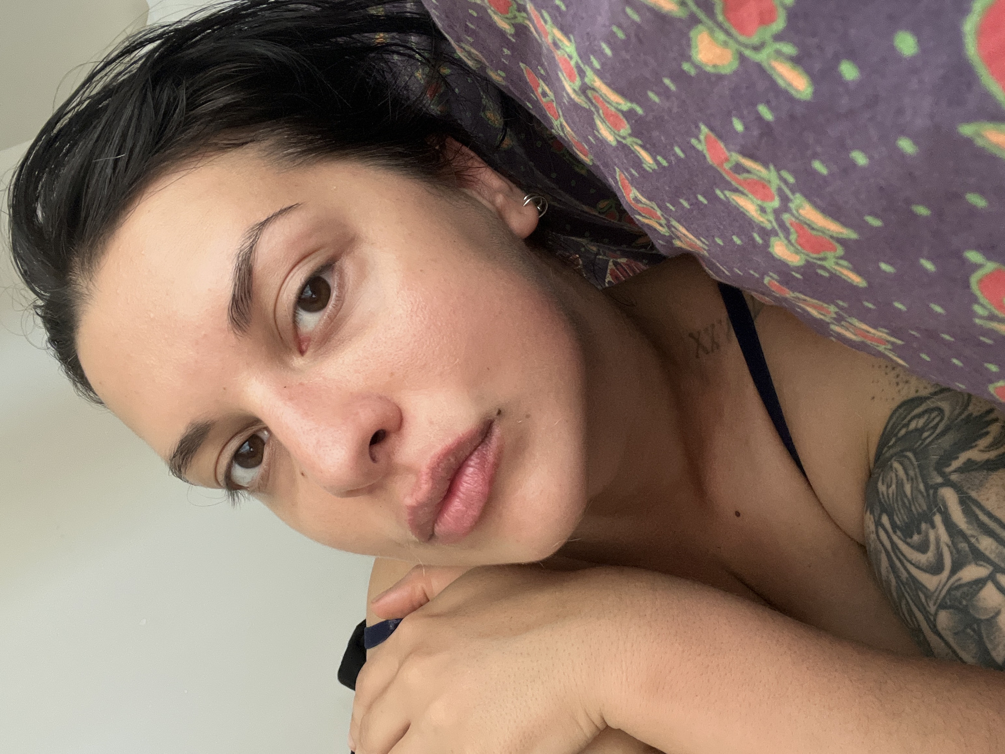 A close-up selfie shows a woman lying on her side, propped up on her arm and resting her head on a pillow. She has dark hair and brown eyes, with a natural makeup look. Her lips are slightly parted, and she is wearing a dark blue top. A large tattoo is visible on her left arm. The pillow has a purple pattern with red and green floral designs. The background is a plain, light-colored wall. There is no indication of the city of Da Nang, Vietnam, or any other specific location.