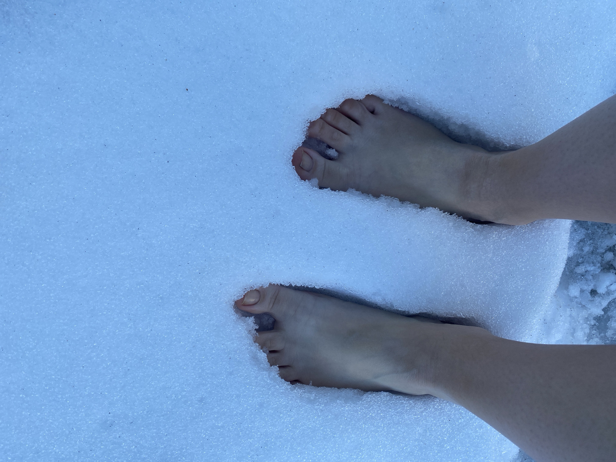 The image shows a pair of bare feet standing in the snow. The feet are pale and have pinkish toenails. The snow is white and covers the ground. The location is not discernible from the image and cannot be identified as Izhevsk, Russia.
