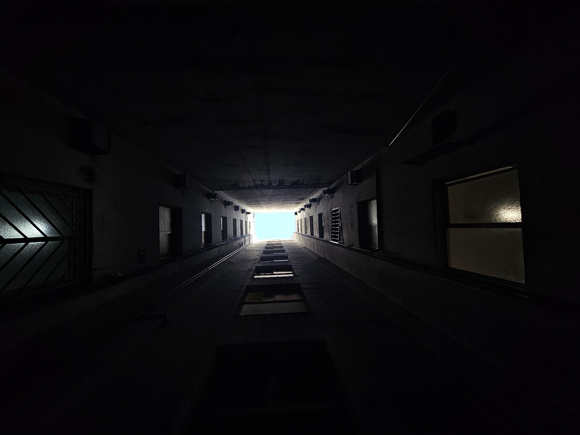 The image shows a low-angle view looking up into a narrow courtyard between tall buildings in Buenos Aires, Argentina. The walls on either side are dark, but show windows and air conditioning units. The sky is visible as a small bright rectangle at the top of the frame. The courtyard appears to be a service area with utility access points visible along the length. The overall impression is one of confinement and darkness punctuated by the narrow slice of bright sky above.