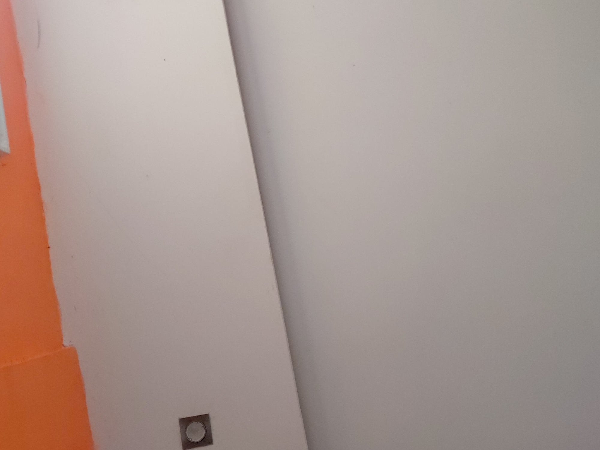 The image displays an abstract view of an indoor architectural space, likely within a building in Santa Ana, Venezuela. The composition is dominated by two large, smooth wall surfaces. The wall on the left features a vibrant orange painted section, sharply contrasting with the larger light gray or off-white wall that covers most of the frame. These two surfaces meet at a distinct diagonal line, suggesting an internal corner or a deliberate architectural feature.
Near the bottom left corner, embedded in the light gray wall, is a small, square-shaped fixture with a metallic or silver finish. It contains a circular element in its center, which could be an indicator light, a button, or a sensor. The walls appear to be painted, with a generally smooth texture, though minor imperfections or specks are visible.
The lighting is even and soft, characteristic of an indoor environment, with no harsh shadows or direct external light sources evident. There are no people, visible activities, or discernible text present in the image. The scene is static, focusing on color contrast and geometric forms within the interior setting.