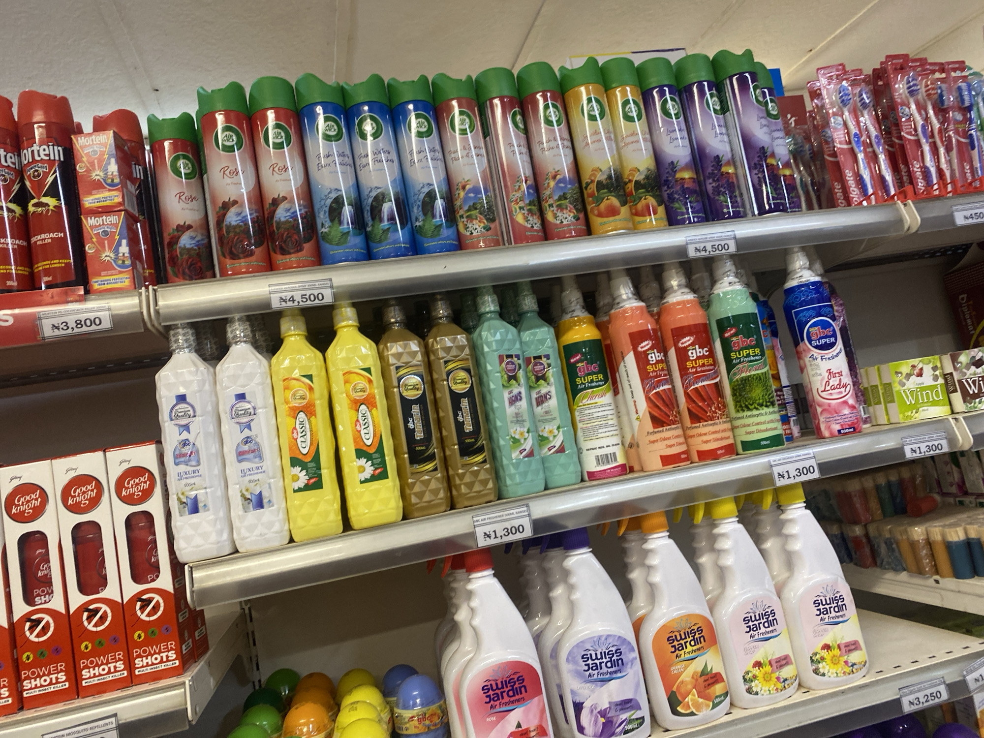 The image shows a retail store shelf displaying a variety of air freshener products and insecticide sprays. The items are arranged in rows on a white metal shelf, with price tags visible. The products include aerosol cans of "Air Wick" in various scents like "Rose," "Fresh Waters," and "Peach & Wild Raspberry," as well as "Mortein Power Shots" insecticide. Below these, there are bottles of "Good Knight Power Shots" and other branded air fresheners such as "Classic," "gbc SUPER Air Freshener," and "Swiss Jardin Air Fresheners" in scents like "Rose" and "Orange." The store appears to be located in Ikorodu, Nigeria, as indicated by the currency symbol "₦" on the price tags and the context of the request. The lighting suggests an indoor environment. There are no people visible in the image.