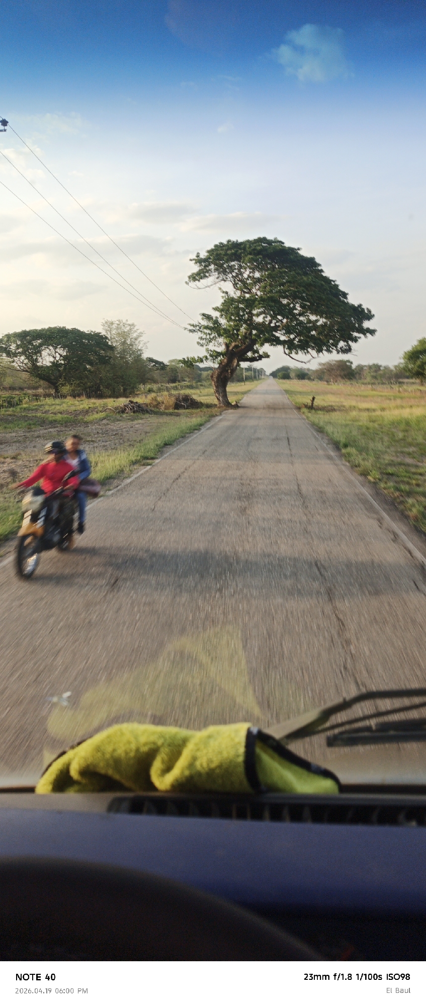 The photograph captures a view from inside a moving vehicle, possibly a car or truck, traveling down a rural road in El Baul, La Cruz, Venezuela. The road stretches into the distance, flanked by fields and trees. Notably, a large tree stands prominently on the right side of the road. A motorbike with two riders is on the left, moving in the same direction as the camera vehicle. The foreground of the image includes the dashboard of the vehicle, with a green towel placed on it. The sky above is clear with blue hues, suggesting daylight. The image appears to have been taken with a wide aperture, as indicated by the EXIF data ("23mm f/1.8"), contributing to the blurred effect due to motion.