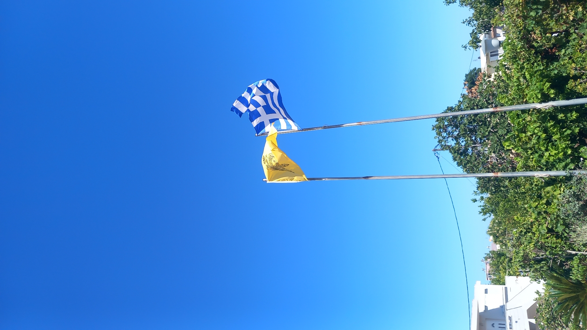 The image shows two flags flying on flagpoles against a clear blue sky. The flag on the right is the national flag of Greece, with its characteristic blue and white stripes and a cross. The flag on the left is yellow and features a black double-headed eagle symbol, likely representing an ecclesiastical or historical Greek flag. Both flags are illuminated by sunlight. Below the flags, lush green trees and foliage are visible, with hints of buildings and structures peeking through the vegetation, suggesting an outdoor setting in a populated area. The overall impression is a sunny day, likely in Greece, given the presence of the Greek flag and the general environment. The location is specified as Platani, Greece.