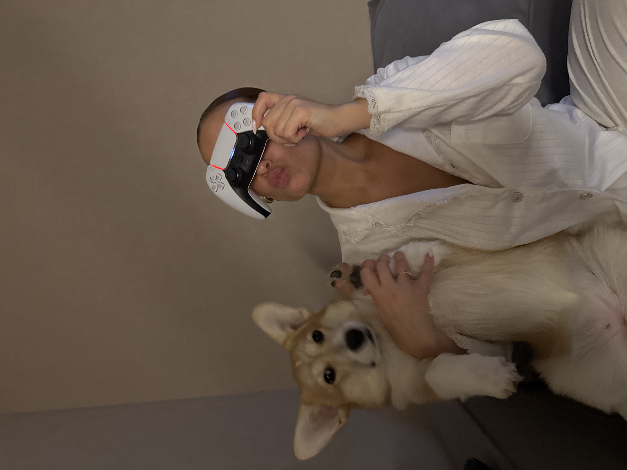 A young woman is sitting on a gray couch, holding a corgi dog. The woman is wearing a white pajama top and is holding a white and black PlayStation 5 controller up to her face like glasses. The dog is on its back, being held by the woman. The scene appears to be indoors, possibly in a home. There is no information provided that allows identification of the city, Chaikovsky, Russia.