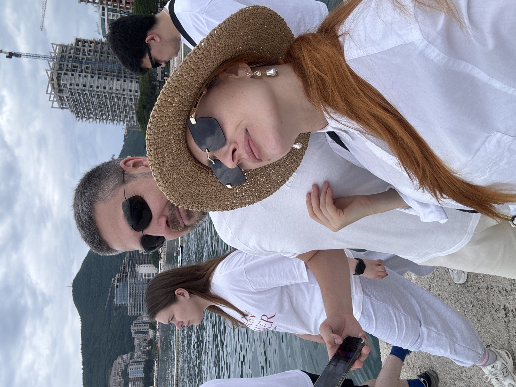 A group of at least four people are posing for a selfie in the city of Budva, Montenegro, on a partly cloudy day. A woman with long red hair wearing a straw hat and sunglasses is taking the selfie. Next to her is a man with gray hair and a mustache, also wearing sunglasses. Behind them is another man in sunglasses and a white shirt. In front of them is a woman with long brown hair looking at a smartphone. All people are wearing white shirts, with two of them also wearing gray pants. The background shows a body of water, buildings, and a mountain. A tall building under construction with a crane is visible behind the first man.