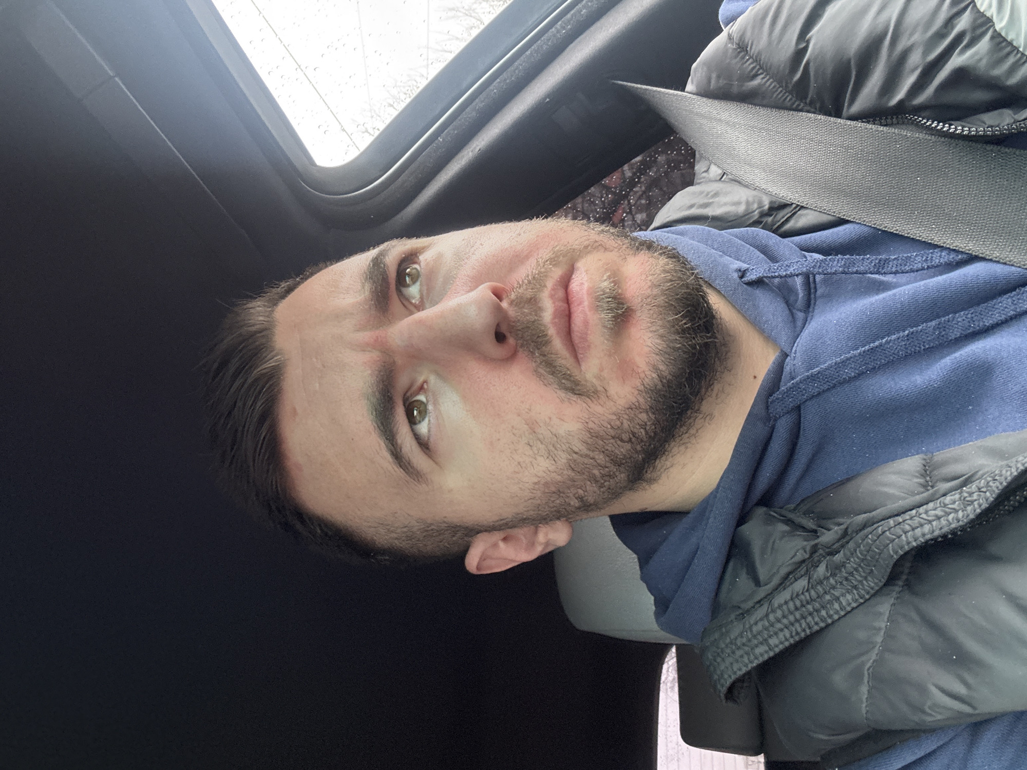 A selfie shot from inside a car shows a man with a beard and light brown hair, wearing a blue hoodie and a black puffer jacket. He is wearing a seatbelt. The car's sunroof is visible with raindrops. The image gives no information about the location in Sofia, Bulgaria.