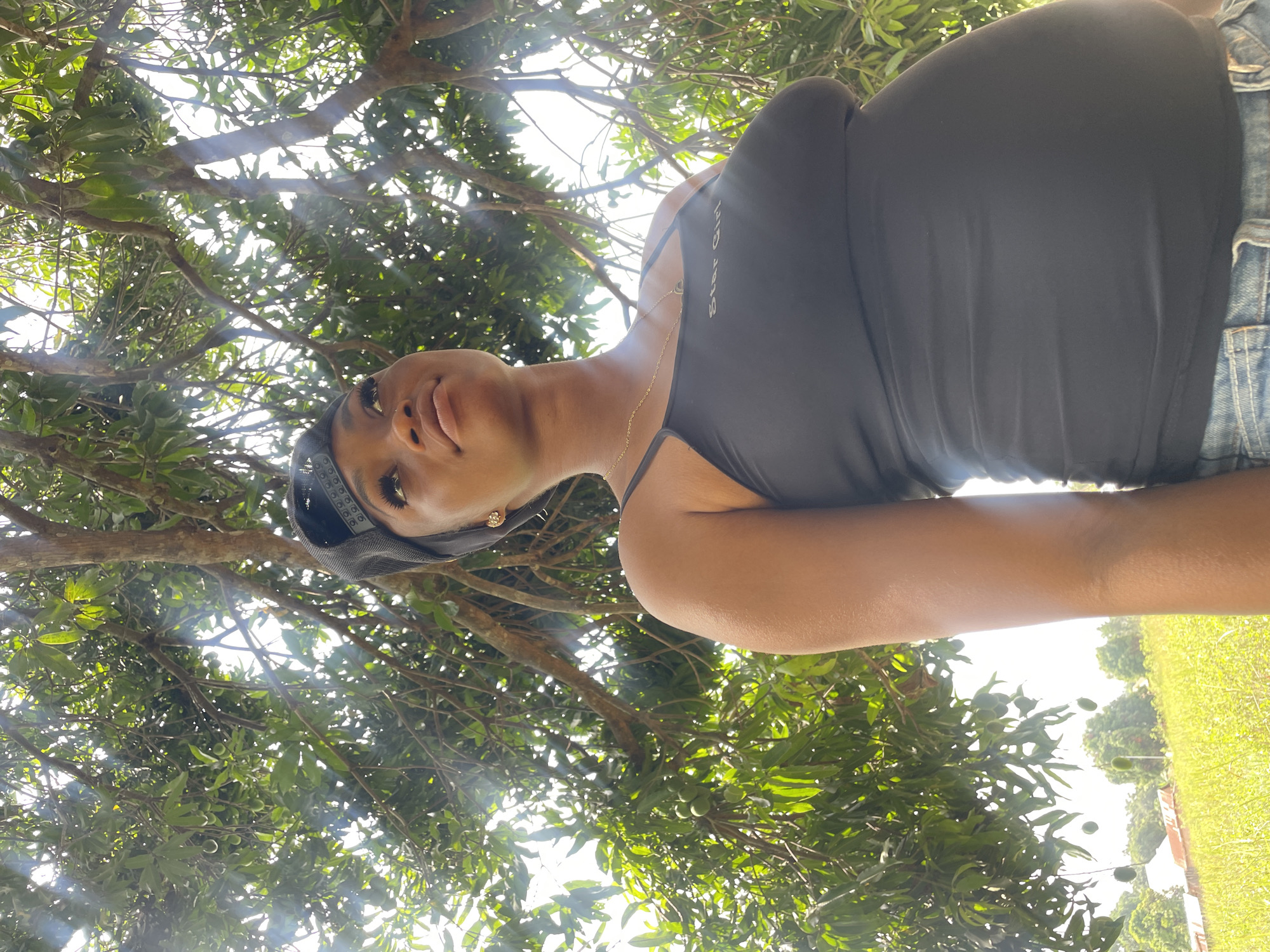 A woman is looking up towards the sky. She is wearing a black tank top, denim shorts, and a black head covering. She has a gold necklace on. The location is outdoors in what looks like a rural area in the city of Ogbe, Nigeria. There are trees with green leaves and green grass in the background. The sky is visible through the trees.