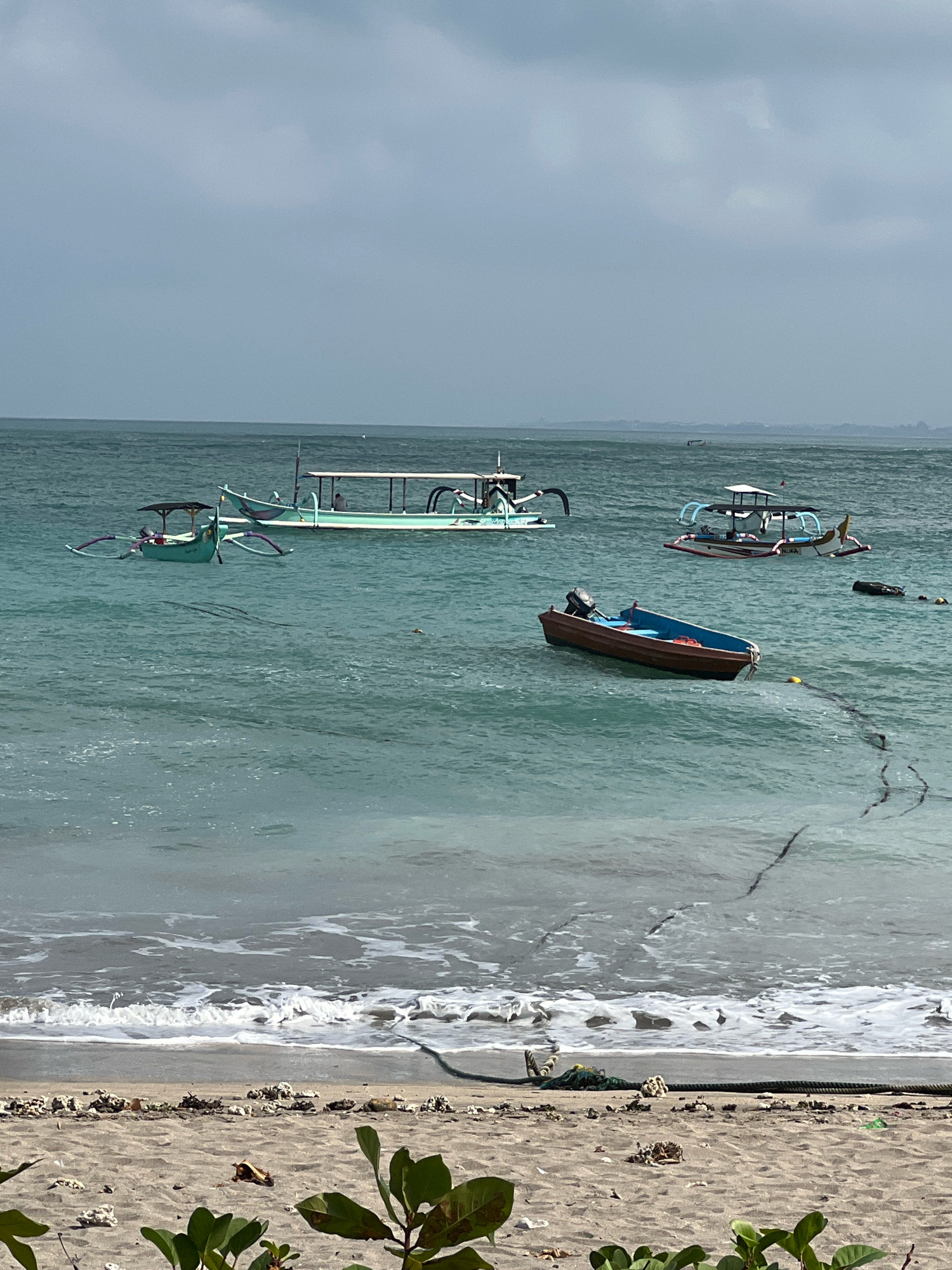 A medium-shot of the ocean with several boats anchored offshore. The water is a turquoise green, with gentle waves lapping at the sandy beach in the foreground. The sky is overcast and a muted gray, suggesting it might be a cloudy day. On the beach, there are scattered pieces of coral and some green foliage in the lower right corner.
In the water, from left to right, there are three traditional Indonesian outrigger boats. The leftmost boat is smaller and painted green, with a dark canopy overhead. The middle boat is larger and pale green, with a white canopy and a person visible inside. The third boat, further to the right, is also an outrigger type, white with a red and white stripe along the hull and a blue canopy. It has the word "NUKA" written on its side.
In front of the "NUKA" boat, a smaller, reddish-brown motorboat is moored. It has a blue interior and an outboard motor at the stern. A dark rope is visible extending from the motorboat towards the shore, disappearing into the water. Several buoys are also visible in the water. In the distance, along the horizon, there appear to be the faint outlines of land, possibly another island or the mainland. The overall scene depicts a typical coastal view in a tropical location. The presence of outrigger boats and the style of the "NUKA" boat suggest a location in Southeast Asia, possibly Indonesia. Given the reference to Kuta, Indonesia, this scene is likely the coast of Kuta.