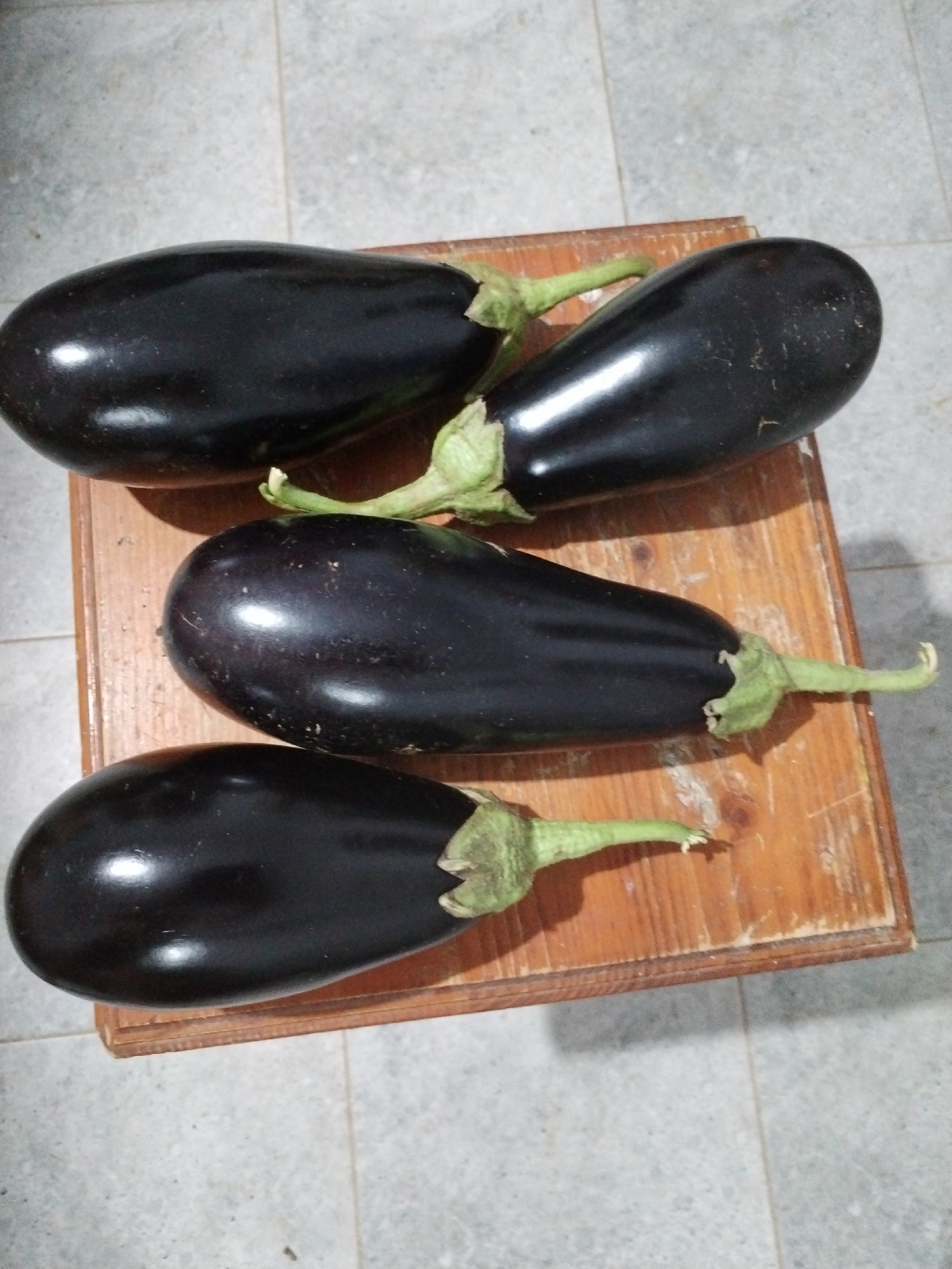 Four large, dark purple eggplants are arranged on a wooden surface. Two eggplants are positioned horizontally, one above the other. The top eggplant is slightly angled with its stem pointing towards the right. Below it, another eggplant lies horizontally with its stem also pointing towards the right. The bottom two eggplants are also placed horizontally and stacked vertically. The uppermost of these two has its stem pointing towards the right, and the lowest eggplant has its stem pointing towards the left. The background consists of a light-colored, tiled floor. The scene is likely indoors, given the lighting and the presence of a wooden surface that could be a table or a stool. The location is specified as Moara Domnească, Romania.