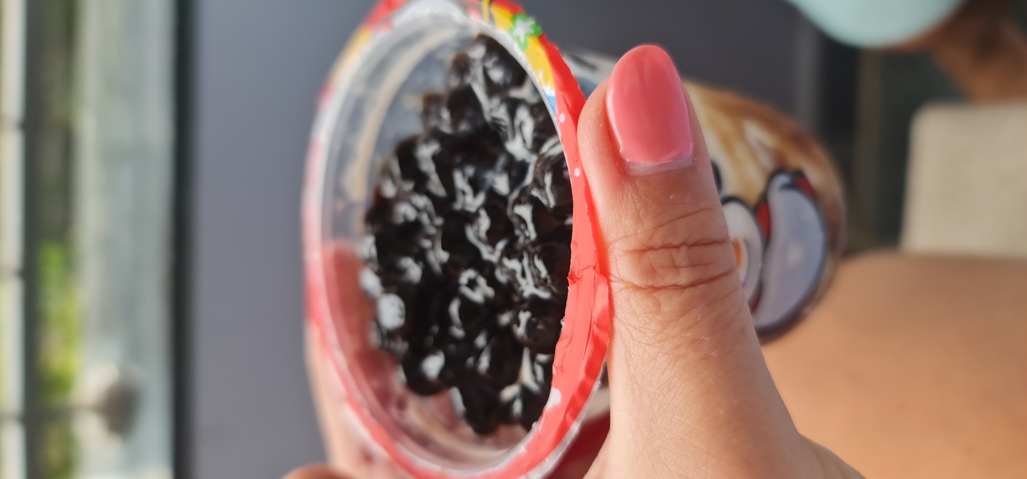 The image captures a close-up view of a person's hand holding a clear plastic cup filled with numerous dark, spherical tapioca pearls, commonly known as boba. The hand, positioned in the lower right and center, has well-maintained nails painted with a light pink polish. The thumb, visible in the foreground, is pressed against the red, colorful rim of the cup, which features abstract patterns in red, yellow, and green. The cup's transparent body allows the dark pearls to be clearly seen against a somewhat blurred background.
Behind the held cup, part of a cartoon-like illustration is faintly visible on the side of another object or the cup itself, showing a white oval with a red element, possibly an eye or cheek. The background is largely out of focus, suggesting a shallow depth of field. To the left, there are blurred vertical elements that could be part of a window frame or structural columns, with hints of green foliage outside or on a distant surface. To the right, the background shows warm, earthy tones, potentially a wall or furniture, and a lighter blue-green object. The lighting suggests it is daytime. No specific location cues are visible within the image to confirm the provided context of Đà Nẵng, Vietnam, nor is any readable text present on the objects. The activity depicted is simply holding the cup of tapioca pearls.