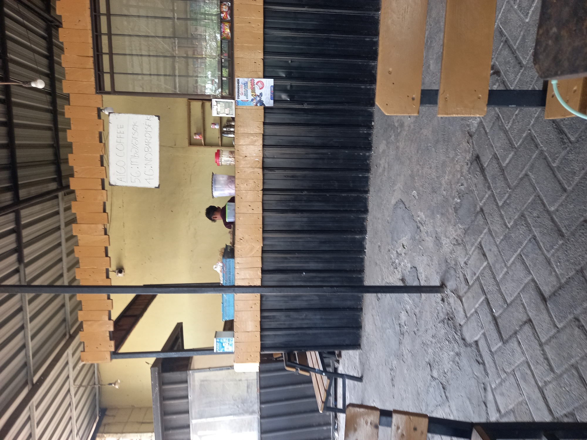 The image depicts an outdoor seating area at what appears to be a food or drink stall in Tunggul, Indonesia. In the background, a counter is set up with shelves holding various items, including drinks, snacks, and a dispenser. A person is partially visible behind the counter, seemingly attending to operations. A sign is posted on the wall above the counter, listing "AICO COFFEE" along with some additional text that is difficult to decipher. Another poster, advertising a "TOURNAMEN" with a prize of "2.000.000," is attached to the front of the counter. The seating area features rustic wooden benches and tables, with a cobblestone-like pavement in the foreground. The overall atmosphere suggests a casual, open-air establishment.