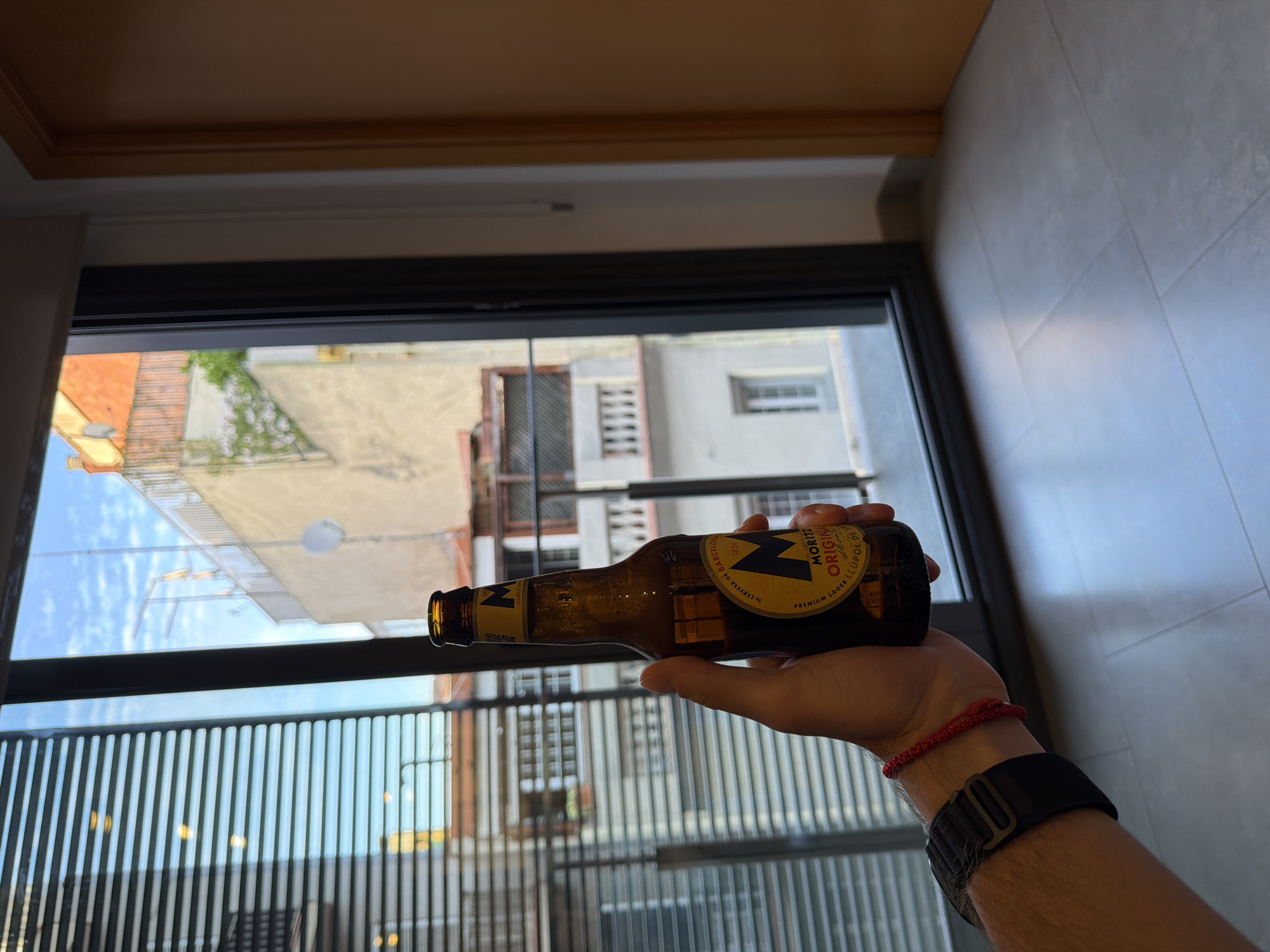 A hand wearing a black watch and a red string bracelet holds a brown glass bottle of Moritz Original beer. The bottle has a yellow label with the letter "M" and text in Spanish, indicating it is from Barcelona, Spain. The hand and bottle are positioned in front of a window with horizontal blinds, through which buildings and a blue sky with clouds are visible. The interior scene includes a tiled floor and a wooden doorframe.
