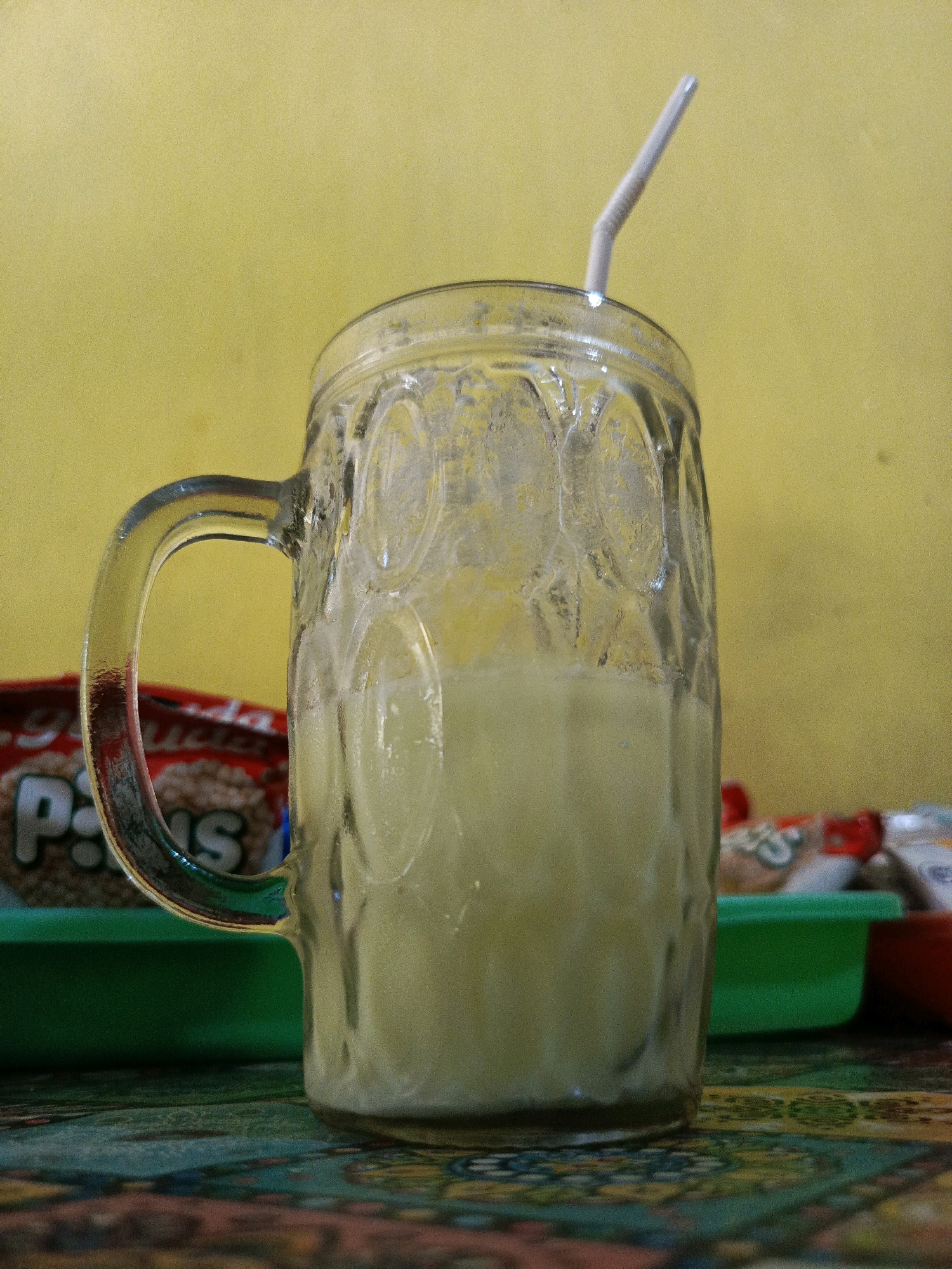 A close-up, eye-level, medium shot captures a clear glass mug filled with a pale yellow beverage, likely juice or a smoothie. The mug has a distinct textured pattern of circular indentations and a curved handle. A white plastic straw with a bent section is inserted into the drink, emerging from the top of the mug. The mug sits on a surface with a colorful, patterned tablecloth or placemat. In the background, a bright yellow wall is visible. To the left and slightly behind the mug, a portion of a red and white package with the text "P S" is discernable, suggesting it may contain snacks. A green plastic container is also present in the mid-ground. The overall lighting is warm and somewhat dim, creating a soft ambiance. This scene likely represents a casual setting, possibly a cafe or home in Pamekasan, Indonesia, where such beverages are commonly enjoyed.