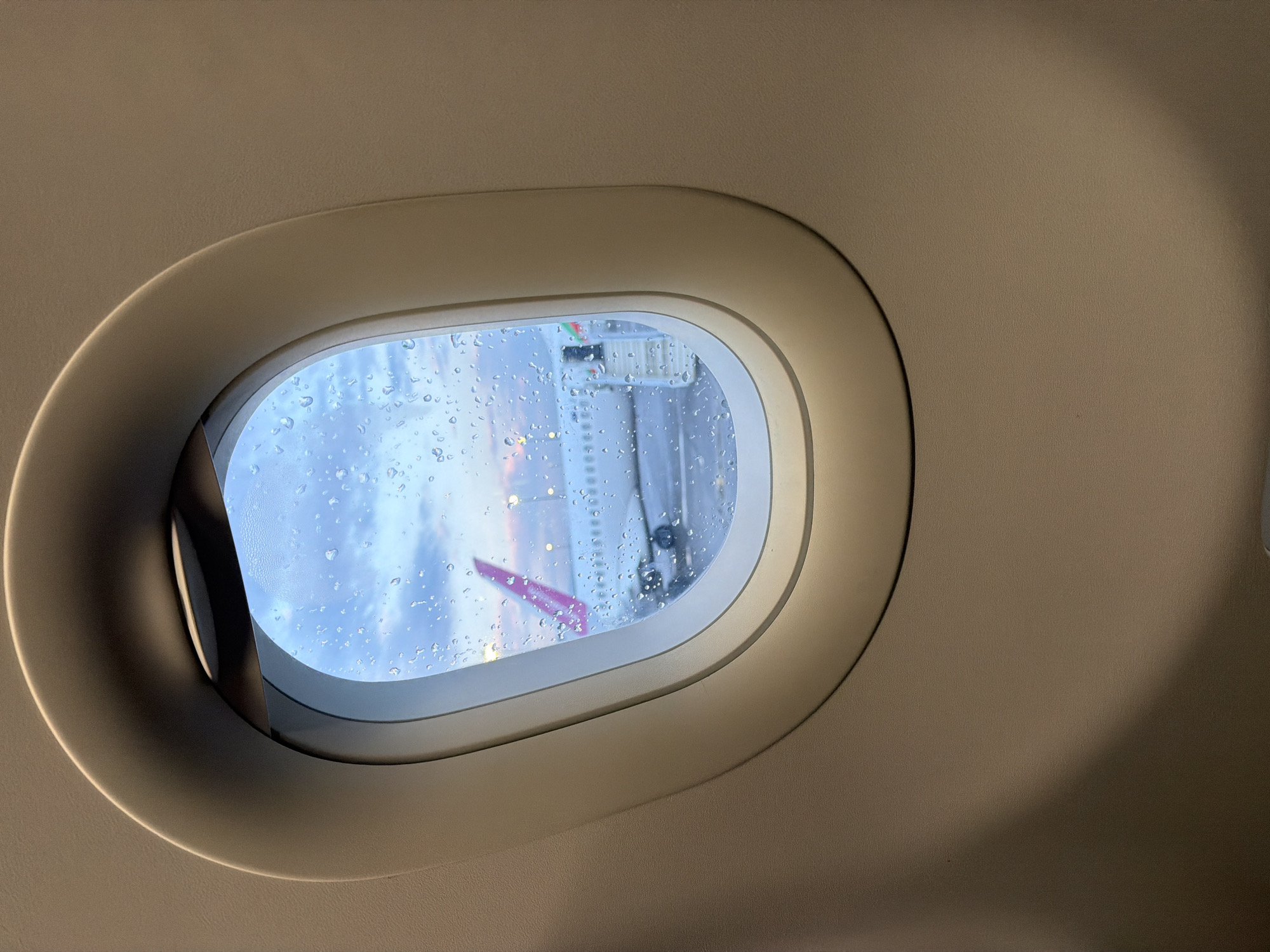 The photograph presents an intimate perspective from inside an airplane cabin, focusing on an oval-shaped window. The robust, light-beige frame of the window contrasts gently with the subtly textured, neutral-toned cabin wall, which features a soft shadow gradient on its right side. The windowpane is covered extensively with numerous water droplets, clearly indicating that it is raining outside.
Through the rain-streaked glass, a blurred yet discernible scene is visible. A substantial portion of a white aircraft structure, possibly a wing or fuselage, occupies the mid-ground. A vibrant magenta or purple-colored detail, potentially part of another airplane's livery or ground equipment, can be seen in the lower left quadrant of the external view. The ambient light beyond the window is diffused and soft, suggesting either early morning, late afternoon, or a heavily overcast day at an airport.
The scene implies the aircraft is stationary on the tarmac, likely preparing for departure or having recently arrived in Sofia, Bulgaria, amidst wet weather conditions. No people are visible within the frame, and there is no legible text present in the image. The overall atmosphere conveyed is one of quiet waiting, characteristic of travel during inclement weather.