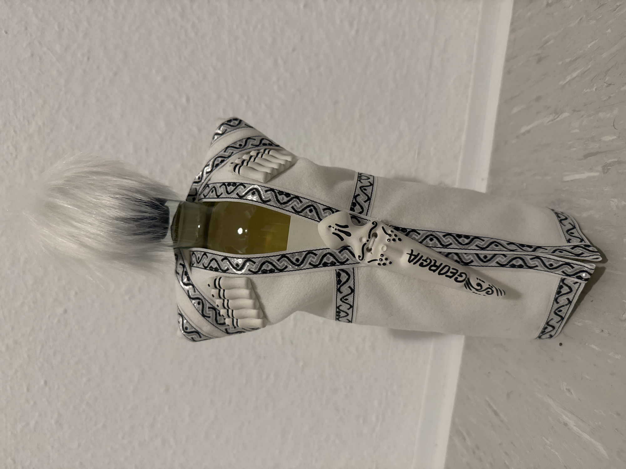 A bottle of wine is dressed in a white fabric resembling a traditional Georgian garment with black and silver trim. The garment has decorative white cylindrical elements on the shoulders, suggestive of cartridges or epaulets, and a decorative dagger-like ornament is attached to the front, inscribed with the word "GEORGIA". The bottle is topped with a fluffy white pom-pom or wig. The object is placed on a light gray countertop against a textured white wall.