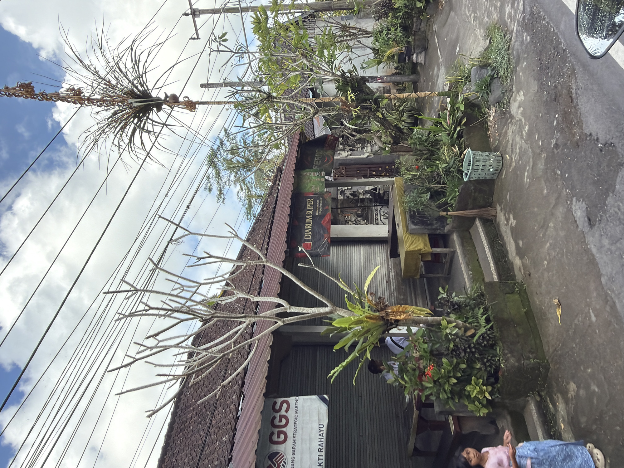 The image depicts a street scene in Tampaksiring, Indonesia, with a storefront and a young girl. The storefront has a corrugated metal roof and roll-up shutters. A banner for "GGS" is visible on the left side of the building, and another banner for "DJARUM SUPER" is on the right. Several potted plants and trees are arranged in front of the storefront. A young girl wearing a pink shirt and a blue skirt stands on the wet pavement on the left side of the image, looking towards the left. Two people are seated at a table inside the storefront. The street is paved with asphalt, and overhead power lines are strung across the sky. A light blue plastic basket and a broom are placed near the entrance of the storefront. The sky is partly cloudy.