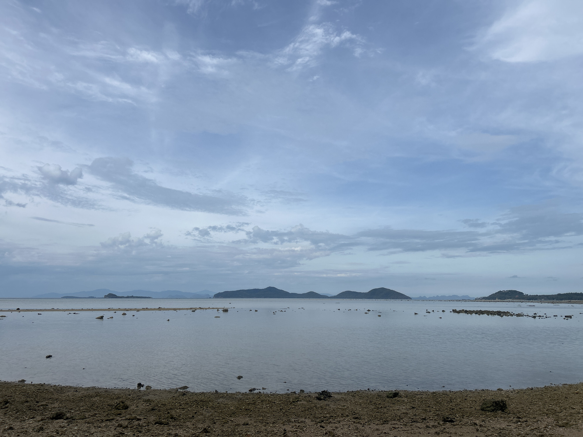 The image depicts a serene coastal scene near Baan Harn, Thailand. The foreground features a rocky, muddy shoreline transitioning into a calm, gray body of water. Scattered rocks are visible both on the shore and within the shallow water. In the distance, several islands with low, rounded hills break the horizon line under a cloudy sky with shades of gray and blue. No people are visible. The environment suggests low tide.