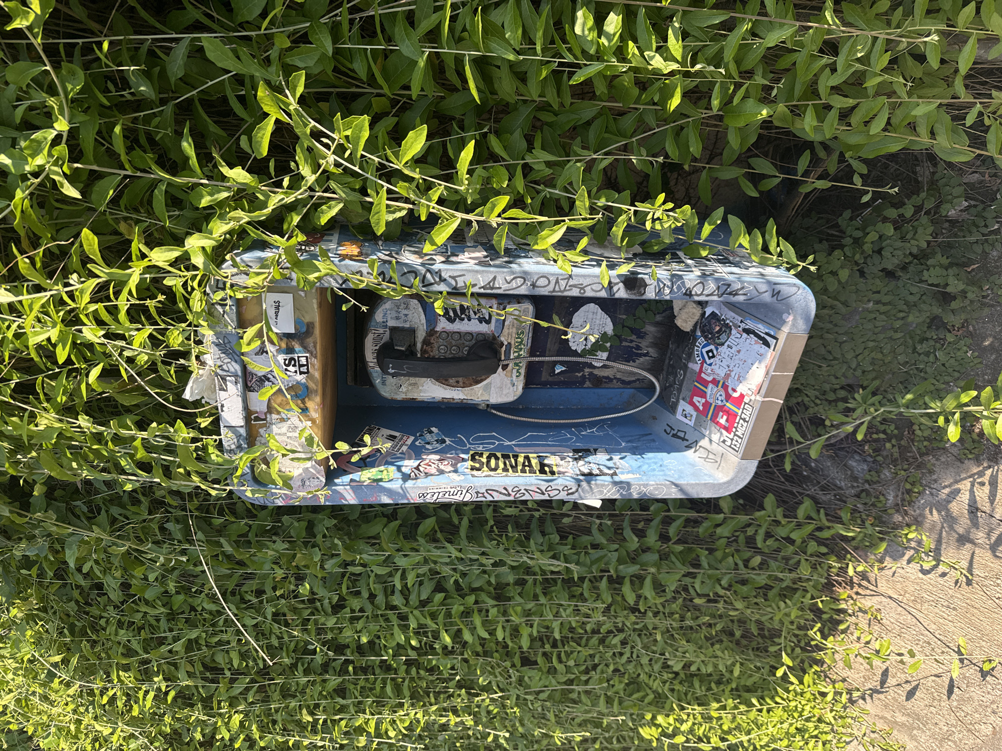 The image depicts an open public telephone box nestled amidst dense green vegetation, likely ground cover or vines. The box itself is aged and covered in various stickers and graffiti, some of which include the words "SONAR," "Timeless," "ROASN3," "MASLIALS," and "HAMBURG." The telephone handset inside is old-fashioned and appears to be out of service. The box is situated outdoors, as evidenced by the surrounding plant life and the presence of concrete or pavement visible in the lower right corner of the frame.
The location is suggested to be in Kuta, Indonesia, but there's no conclusive evidence within the frame to verify the geographic location. The presence of the vegetation and the deteriorated condition of the telephone box indicate a humid climate and possibly a neglected or underutilized infrastructure element.