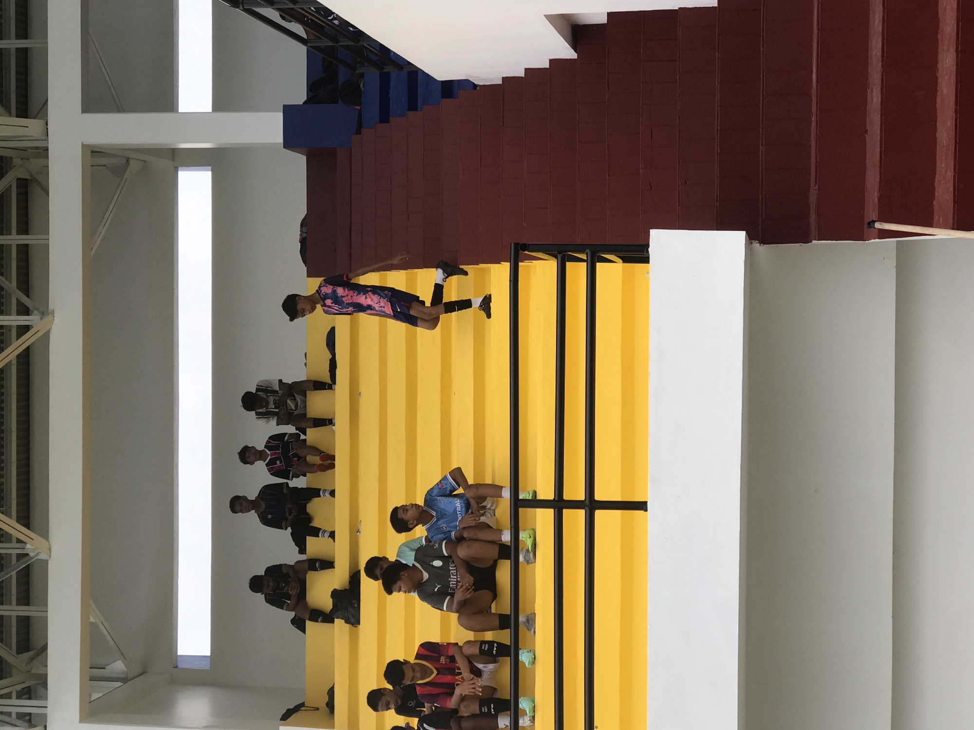 The image shows a group of young people sitting on tiered seating, seemingly in an indoor stadium or auditorium in Palohlada, Indonesia. The tiered seating is painted yellow, with the upper tiers painted burgundy and blue. A boy wearing a pink and blue sports jersey stands on an upper tier. The seating is partially obscured by a black metal railing and a white pillar. The walls are white, and a structure with vertical bars is visible on the left side of the image.